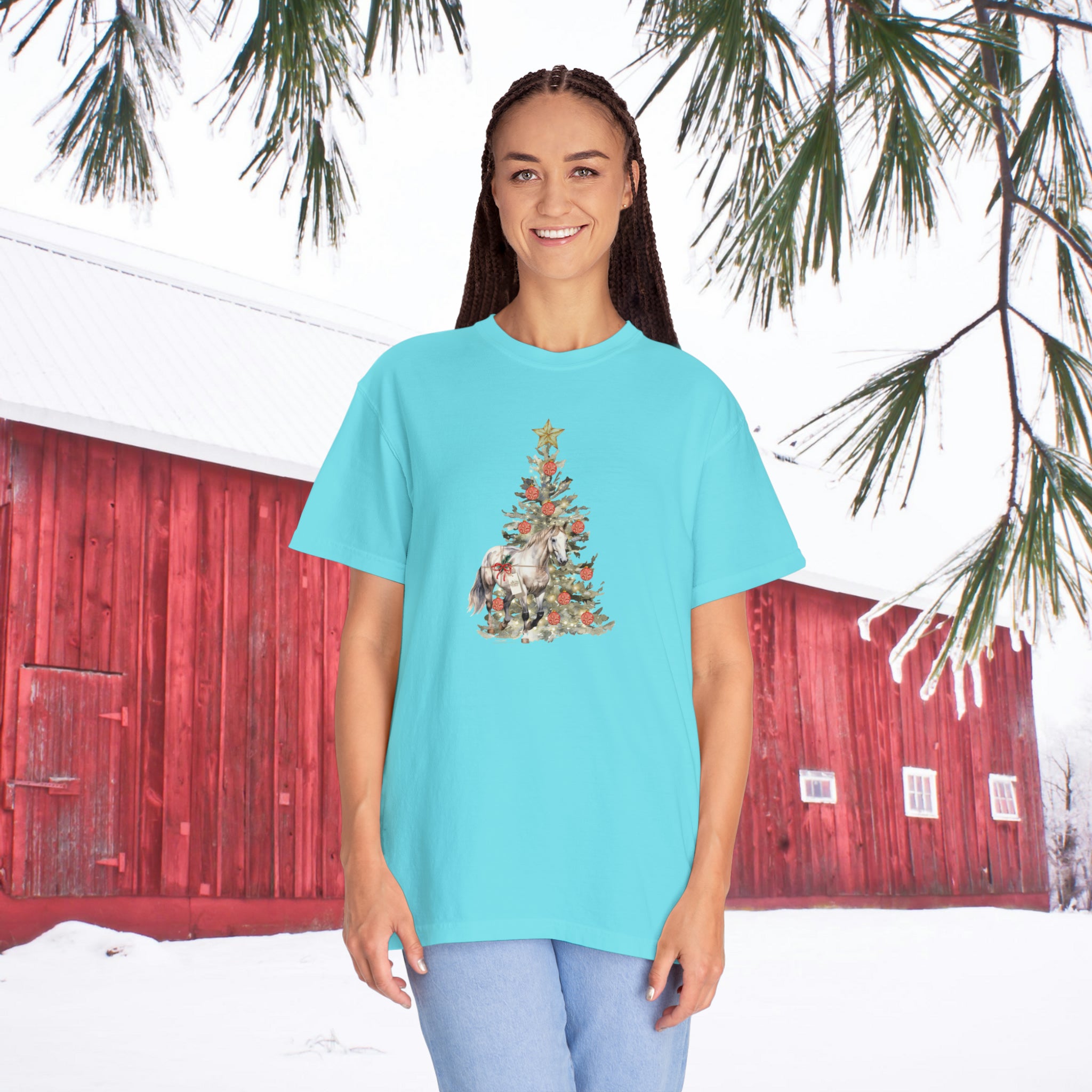 A person wearing "The Gift of A Horse" Christmas garment-dyed t-shirt by Urban SoulShine & co, featuring a light blue shade and ring-spun cotton, stands in front of a red barn. The ground and barn roof are covered in snow, while pine branches hang overhead, creating a picturesque winter scene.