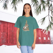 A person stands smiling in front of a red barn with snow on the ground and evergreen branches above. They are wearing The Gift of A Horse Christmas Garment-Dyed T-shirt by Urban SoulShine & Co, made of soft ring-spun cotton and featuring a festive Christmas tree design, paired with light blue jeans.