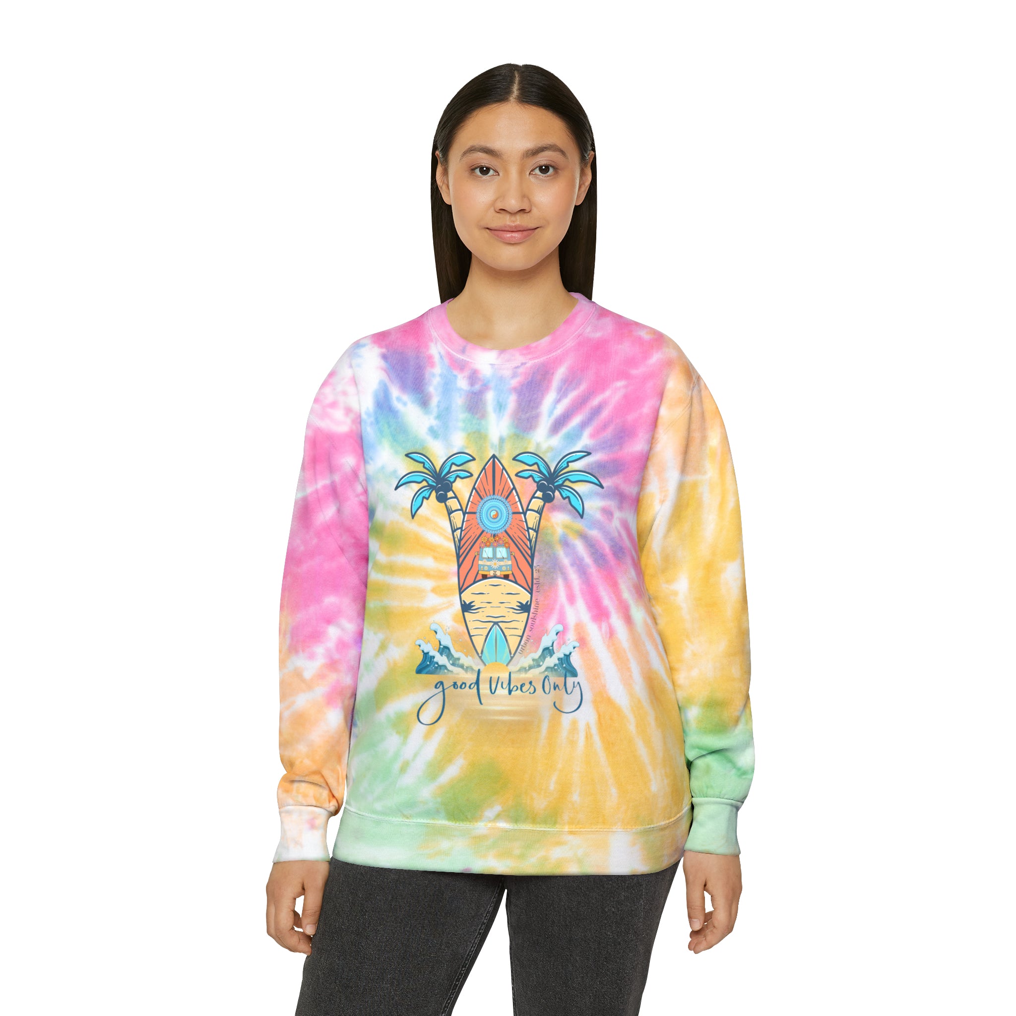 A woman is standing and facing the camera, wearing an Urban SoulShine & co Surf's Up Good Vibes Only Tie-Dye Sweatshirt (unisex) made of pre-shrunk PFD cotton. The sweatshirt features an illustration of a surfboard, palm trees, and waves, with the text "Good Vibes Only" beneath the design. She has long, dark hair and is smiling slightly.