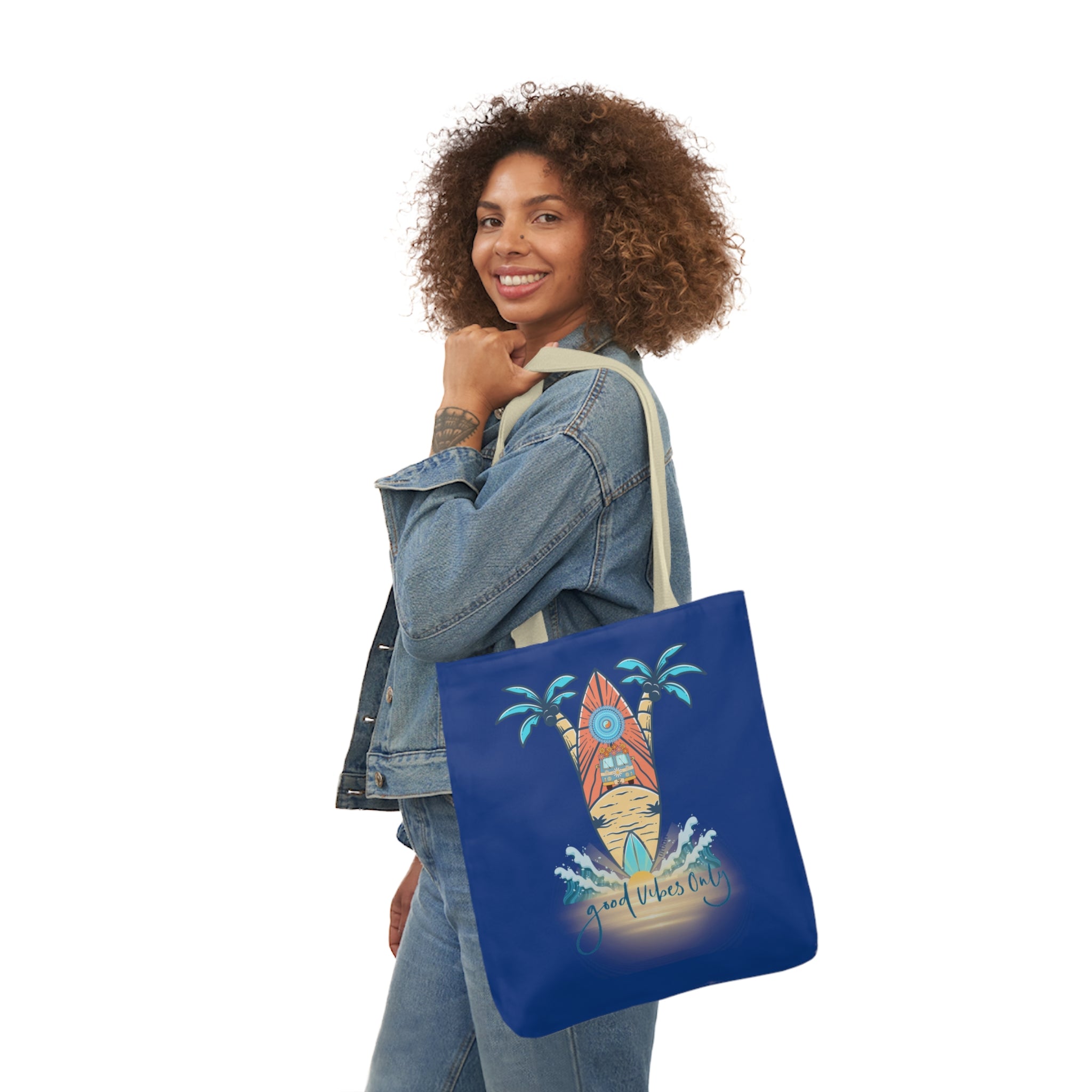 A person with curly hair, wearing a denim jacket, is smiling at the camera. They hold a blue tote bag on their shoulder. The tote bag features a surfboard design with palm trees and the text "Good Vibes Only," reflecting their artistic expression and soulshine. This stylish accessory is the Surf's Up Good Vibes Only Beach Tote by Printify.