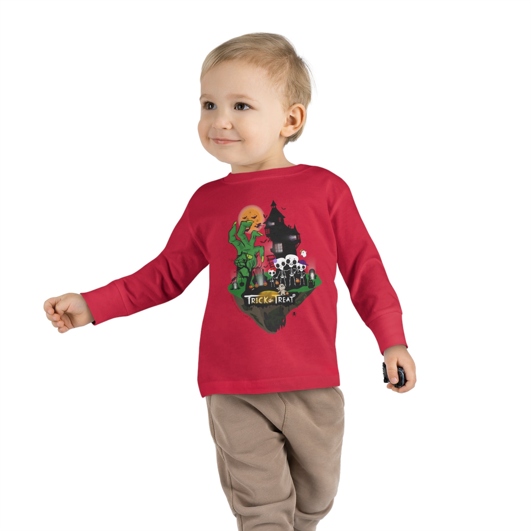 A young child with a slight smile stands against a plain background, wearing a gray long-sleeve shirt and blue jeans. The Spooky Island Halloween Toddler Long Sleeve Tee by Printify features a green, pumpkin-headed figure, a haunted house, and the phrase "Trick or Treat," showcasing an artistic expression perfect for the season.