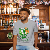 A man with curly hair and a beard sits in front of a bar counter, smiling. He's wearing a grey T-shirt with a colorful graphic of a leprechaun and text that reads "ShamRock Grill" and "McFizzle's Irish PUB" alongside a shamrock and musical notes. His Printify Shamrock Grill & McFizzie's Irish Pub SoftStyle Tee (Unisex) radiates soulshine, embodying the spirit of inspirational wear.