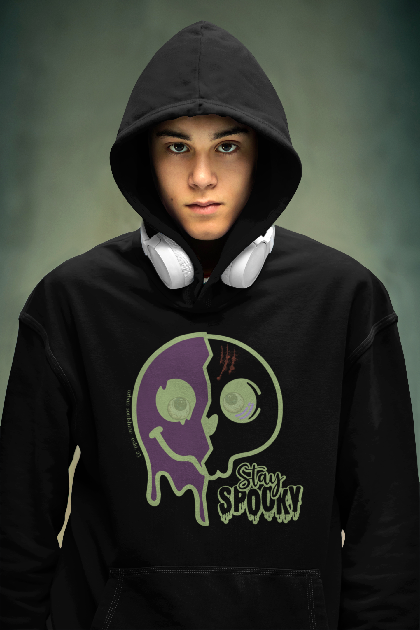 A person wearing a black Stay Spooky Funky Faces Halloween Hoodie (unisex) by Printify with the hood up, featuring a graphic of a dripping, two-faced smiley and the text "Stay Spooky," is showcasing their unique style. White headphones rest around their neck. The background is blurred, displaying muted shades of green and gray—a true testament to how you can express yourself through inspirational wear.