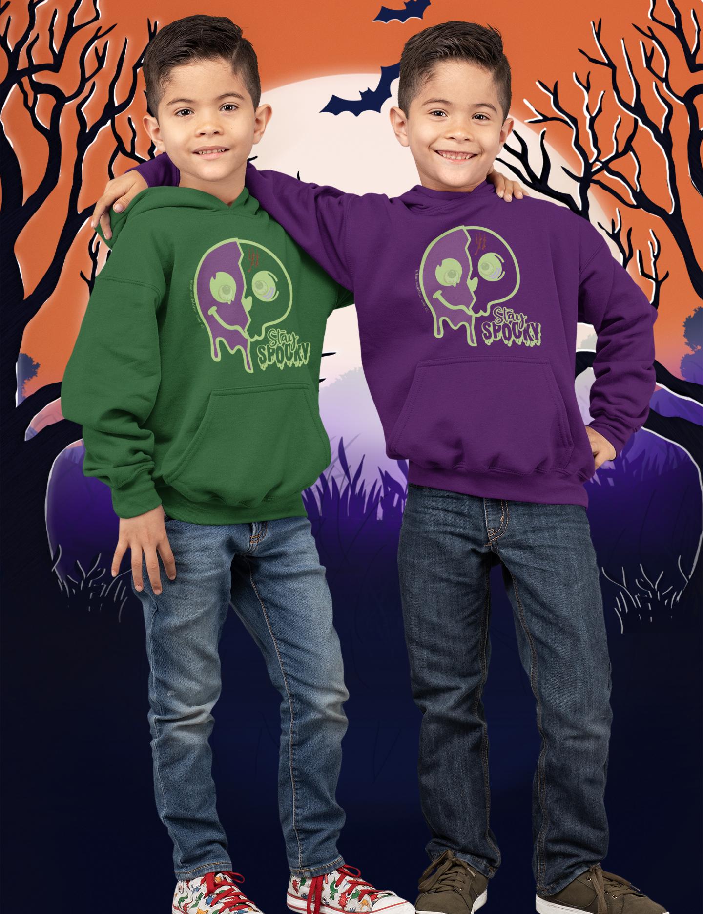 Two young boys stand smiling in front of a Halloween-themed background featuring a full moon, bats, and leafless trees. They are dressed in Printify's Stay Spooky Funky Faces Halloween Youth Hoodie; one wears green, the other purple. Both boys don jeans and sneakers from our soulshine inspirational wear collection.