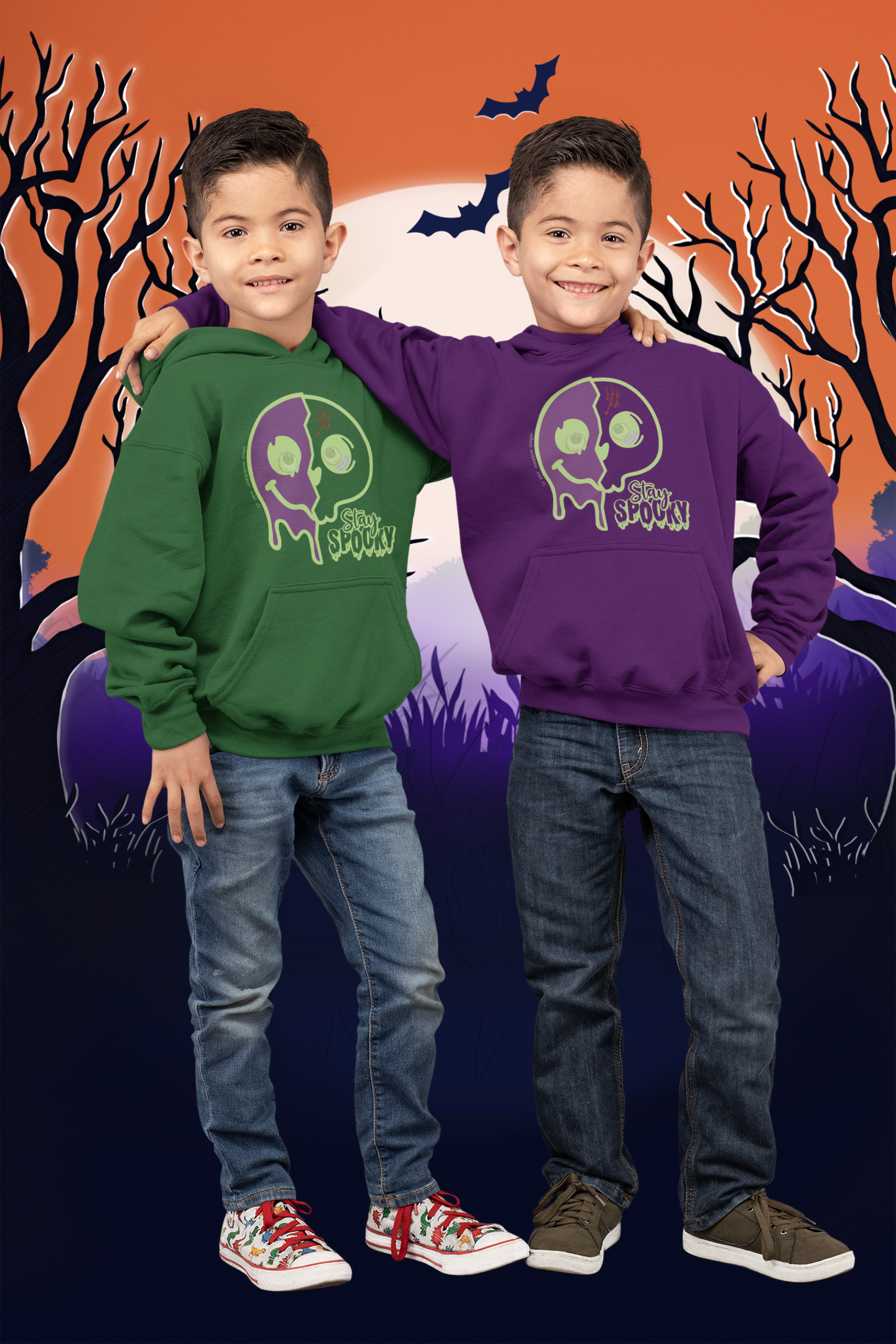 Two young boys stand smiling in front of a Halloween-themed background featuring a full moon, bats, and leafless trees. They are dressed in Printify's Stay Spooky Funky Faces Halloween Youth Hoodie; one wears green, the other purple. Both boys don jeans and sneakers from our soulshine inspirational wear collection.