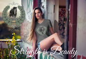 A woman with long hair wearing a gray Urban SoulShine & co Strength is Beauty Horse Tee featuring a horse design is sitting in a partially ruined building. Surrounding the horse image is a leafy wreath. "Strength is Beauty" is written at the bottom in flowing script, daring to inspire. The same horse logo appears on the wall.