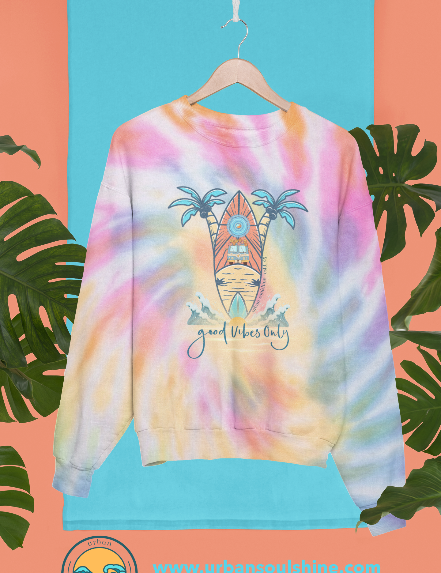 A Surf's Up Good Vibes Only Tie-Dye Sweatshirt (unisex) with the design of a tropical scene, featuring a surfboard, palm trees, and a sun on a beach. The text below the design reads "Good Vibes Only." Made from pre-shrunk PFD cotton, this tie-dye sweatshirt by Urban SoulShine & co is displayed hanging against a blue backdrop with tropical leaves.