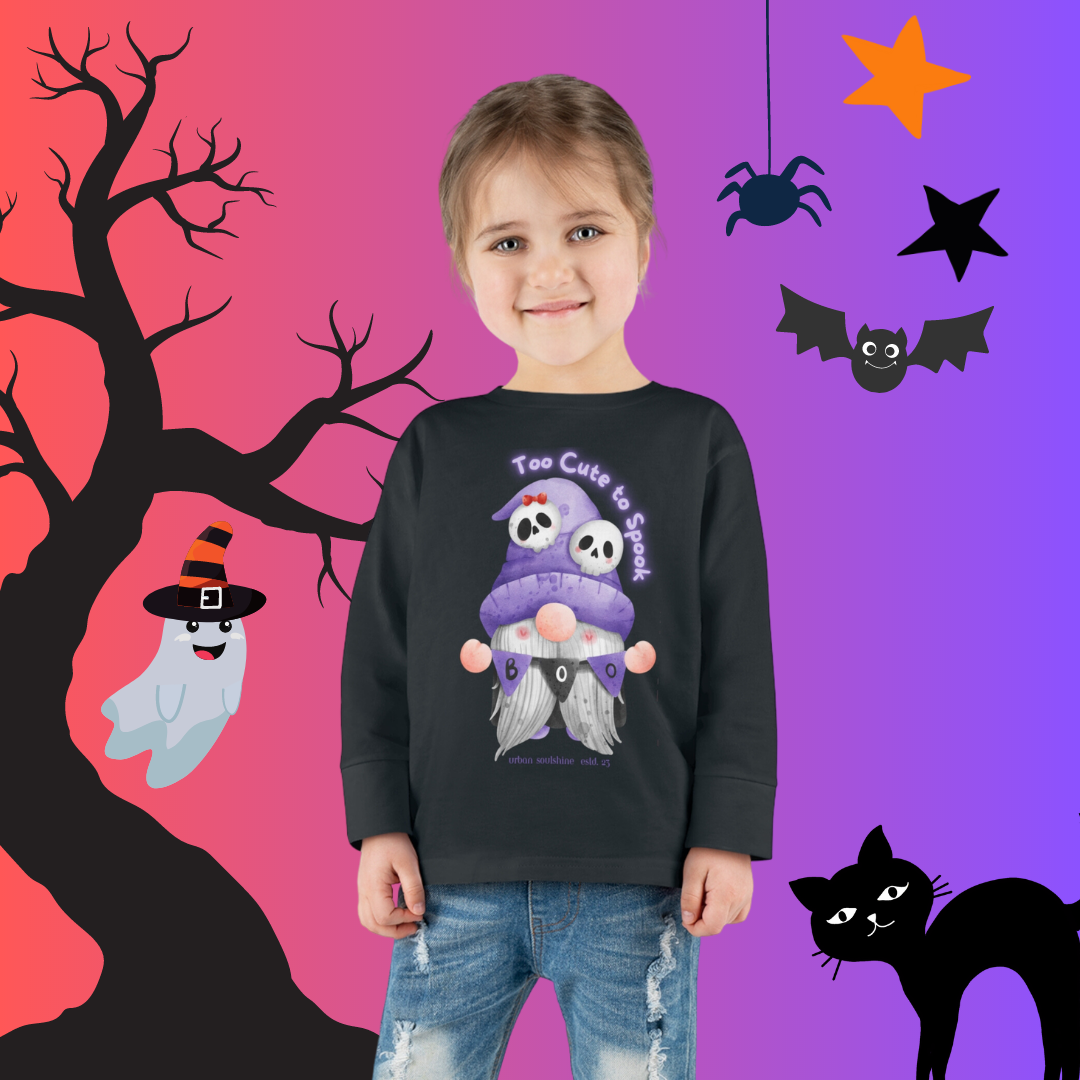 A young child with a ponytail is smiling and wearing a black sweatshirt that says "Too Cute to Spook" with cartoon gnomes in Halloween attire. The background is a colorful gradient with Halloween-themed decorations like a ghost, spider, bat, black cat, and bare tree, showcasing vibrant artistic expression. The child is dressed in the Printify Too Cute To Spook Halloween Toddler Long Sleeve Tee (unisex Tot).