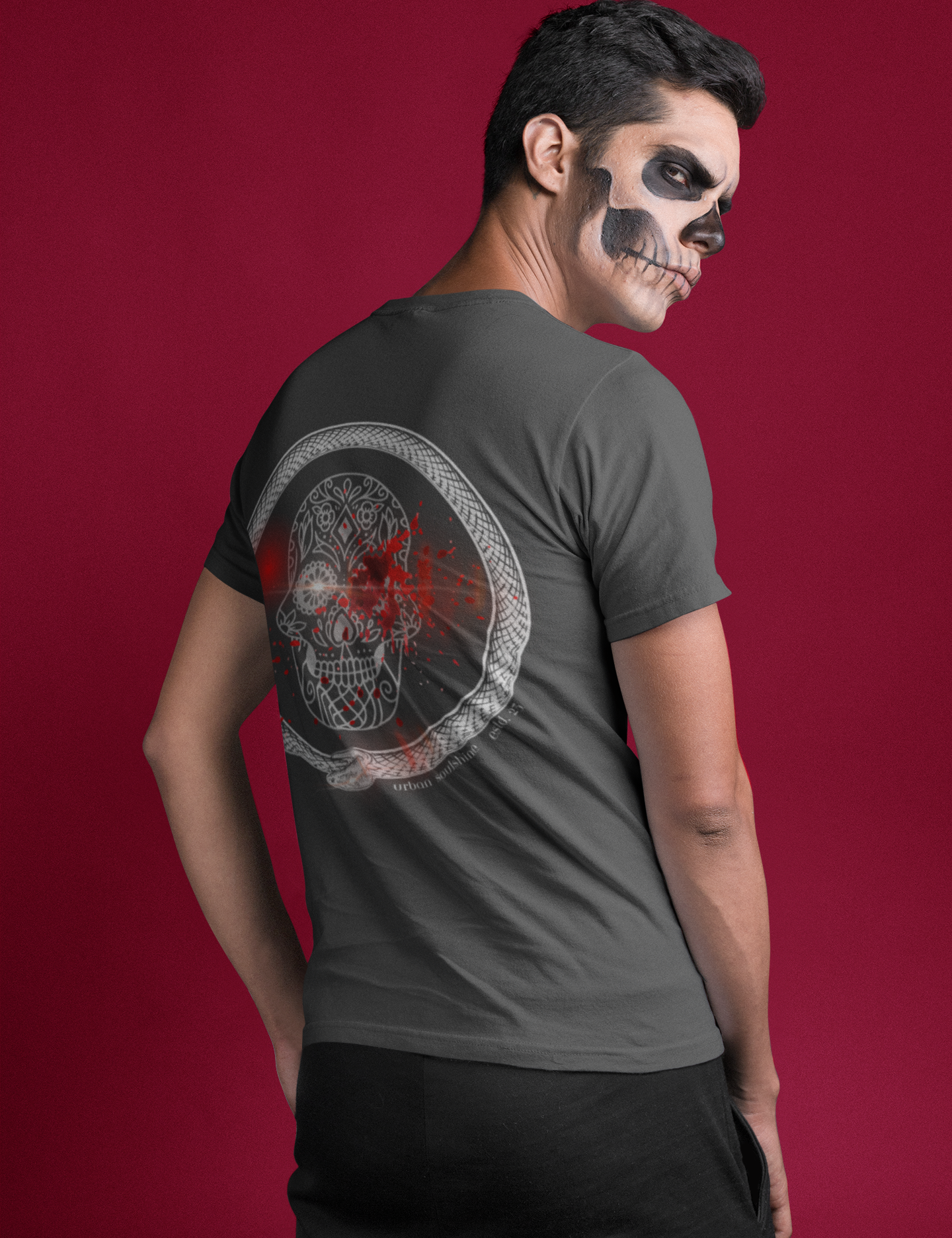 A person with face paint resembling a skull stands against a red background. They are wearing an Urban SoulShine & co Serpent Splatter Skull Tee (unisex) with a graphic of a skull decorated with red and white elements on the back. The person is looking over their shoulder at the camera.