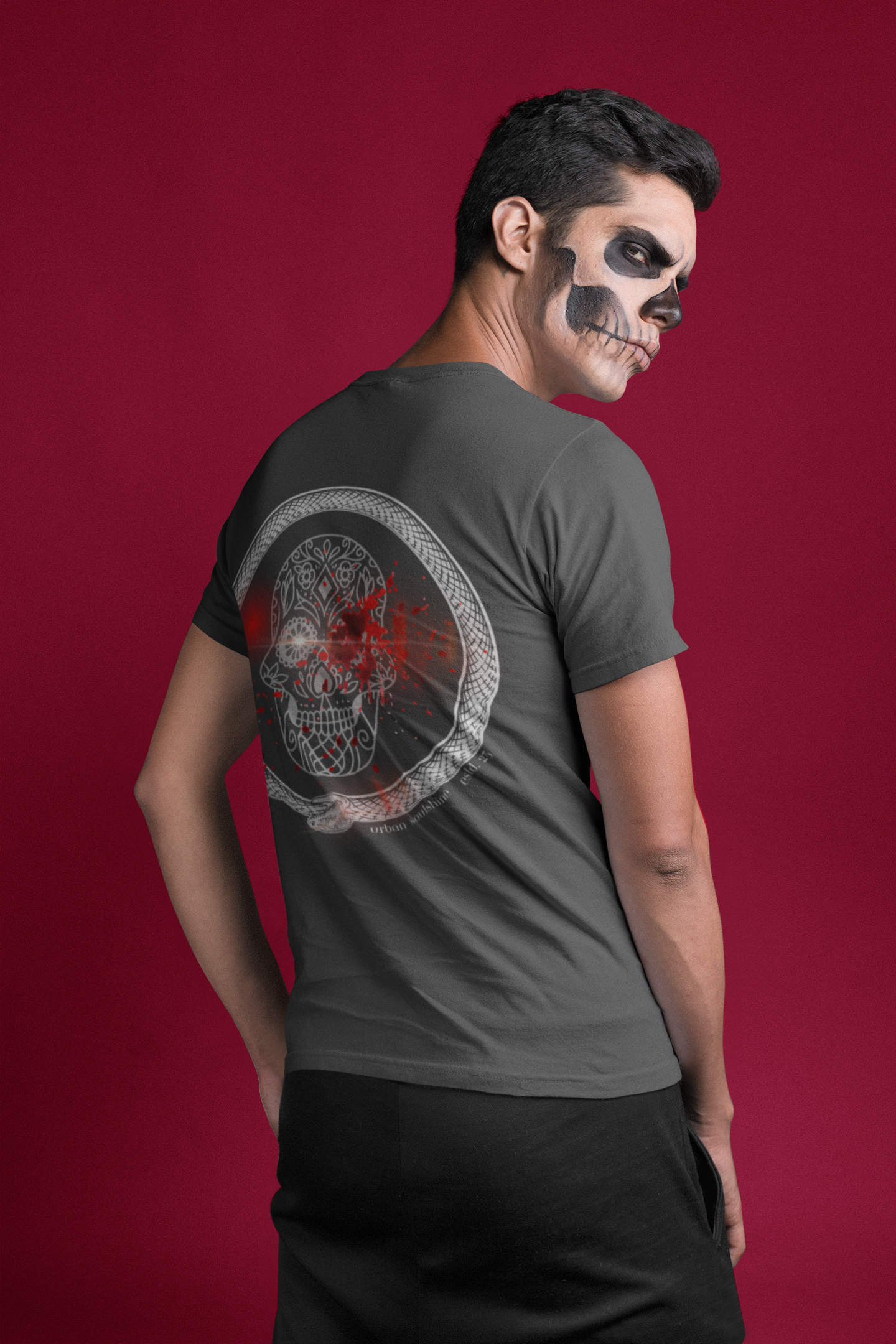 A person with face paint resembling a skull stands against a red background. They are wearing an Urban SoulShine & co Serpent Splatter Skull Tee (unisex) with a graphic of a skull decorated with red and white elements on the back. The person is looking over their shoulder at the camera.