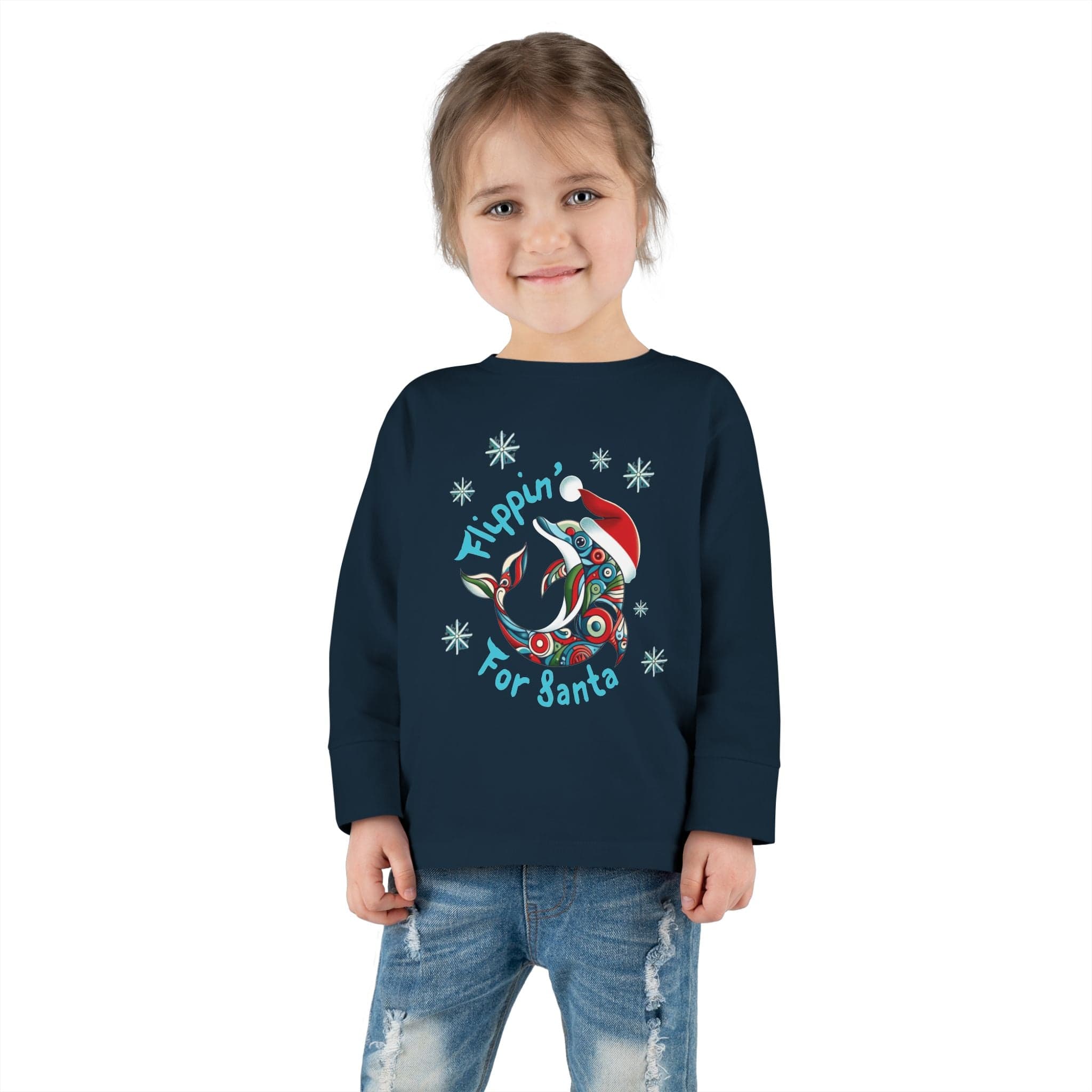 A young child is beaming in the Urban SoulShine & co Flippin' For Santa Christmas Dolphin Long Sleeve Toddler Tee, made of 100% cotton and featuring a festive dolphin design. The cozy unisex fit pairs perfectly with blue jeans against a plain white background.