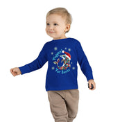 A light-haired young child smiles to the side, wearing the Urban SoulShine & Co Flippin' For Santa Christmas Dolphin Long Sleeve Toddler Tee, a unisex fit made from 100% cotton. The blue tee showcases a festive surfing Santa graphic alongside the "Flippin' for Santa" text. The child has one arm slightly raised and is dressed in beige pants.