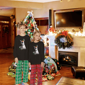 Two children stand in a festive living room decorated for Christmas, wearing matching Merry Donkey Long Sleeve Christmas Pajama Sets (Youth) from Urban SoulShine & Co. These pajamas, with their delightful 100% cotton design, feature a charming reindeer motif. In the background, a Christmas tree illuminated with lights and adorned with ornaments is surrounded by wrapped gifts, while a cozy fireplace adds warmth to the scene.