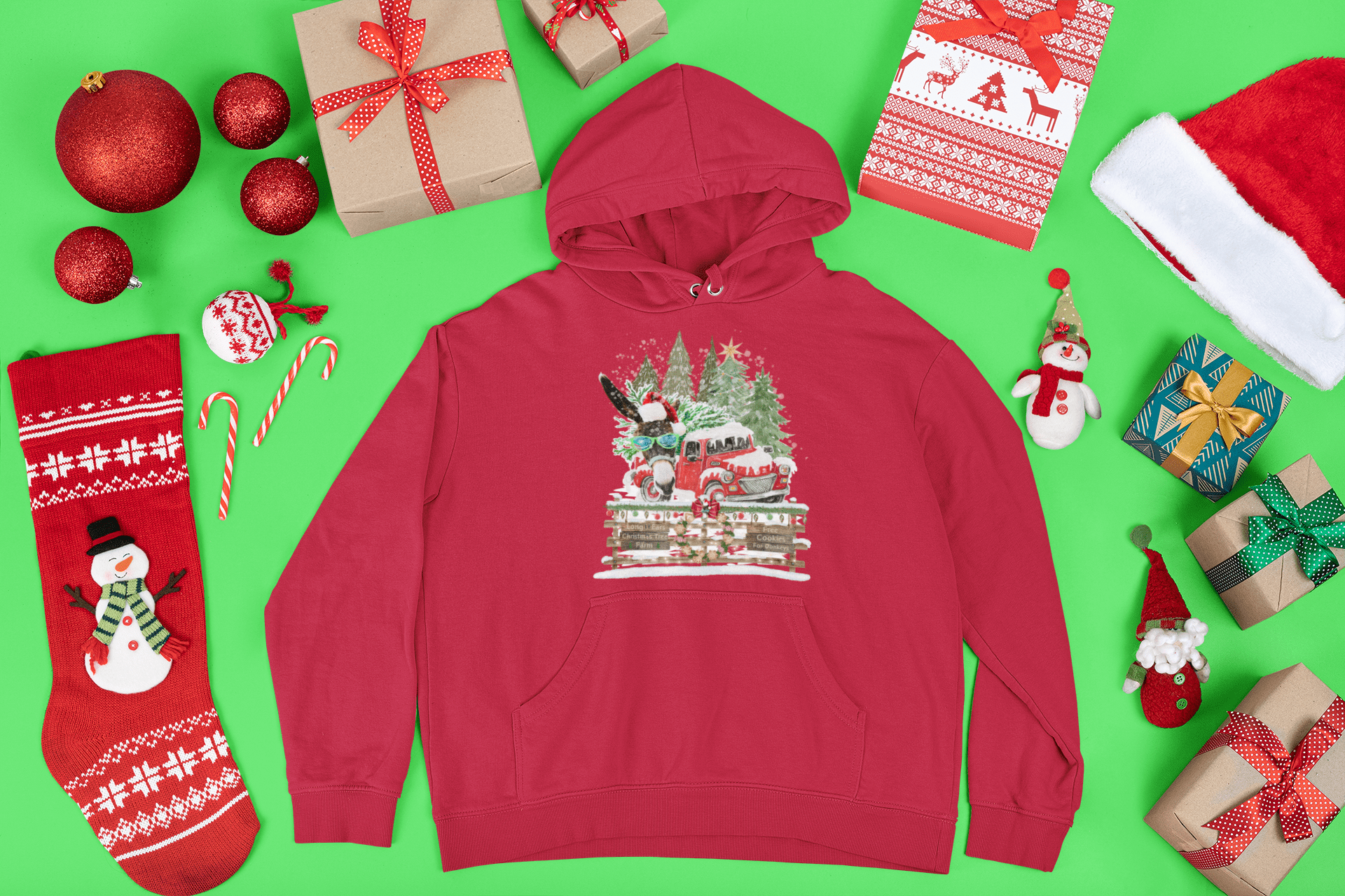 A Long Ears Christmas Tree Farm Donkey Claus Hoodie in red, by Urban SoulShine & co, exudes a cozy feel as it rests on a green background. This unisex hoodie is surrounded by festive items such as wrapped gifts, a Santa hat, Christmas stockings, ornaments, and candy canes.