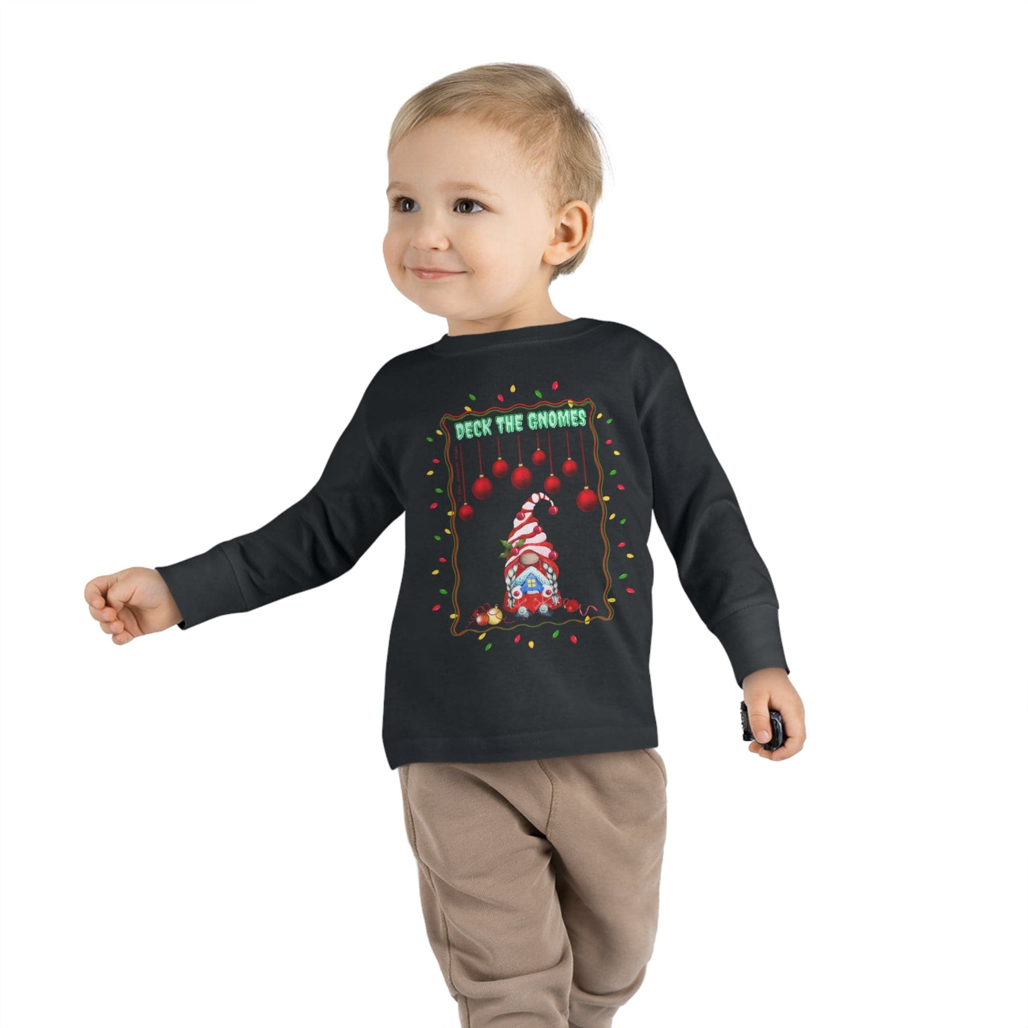 A young child is walking and smiling, wearing the "Deck The Gnomes Christmas Toddler Long Sleeve Tee" by Urban SoulShine & co. This cozy black shirt features a festive holiday gnome design with red and white decorations and the playful text "Deck the Gnomes." Paired perfectly with beige pants, the outfit stands out against a plain white background.