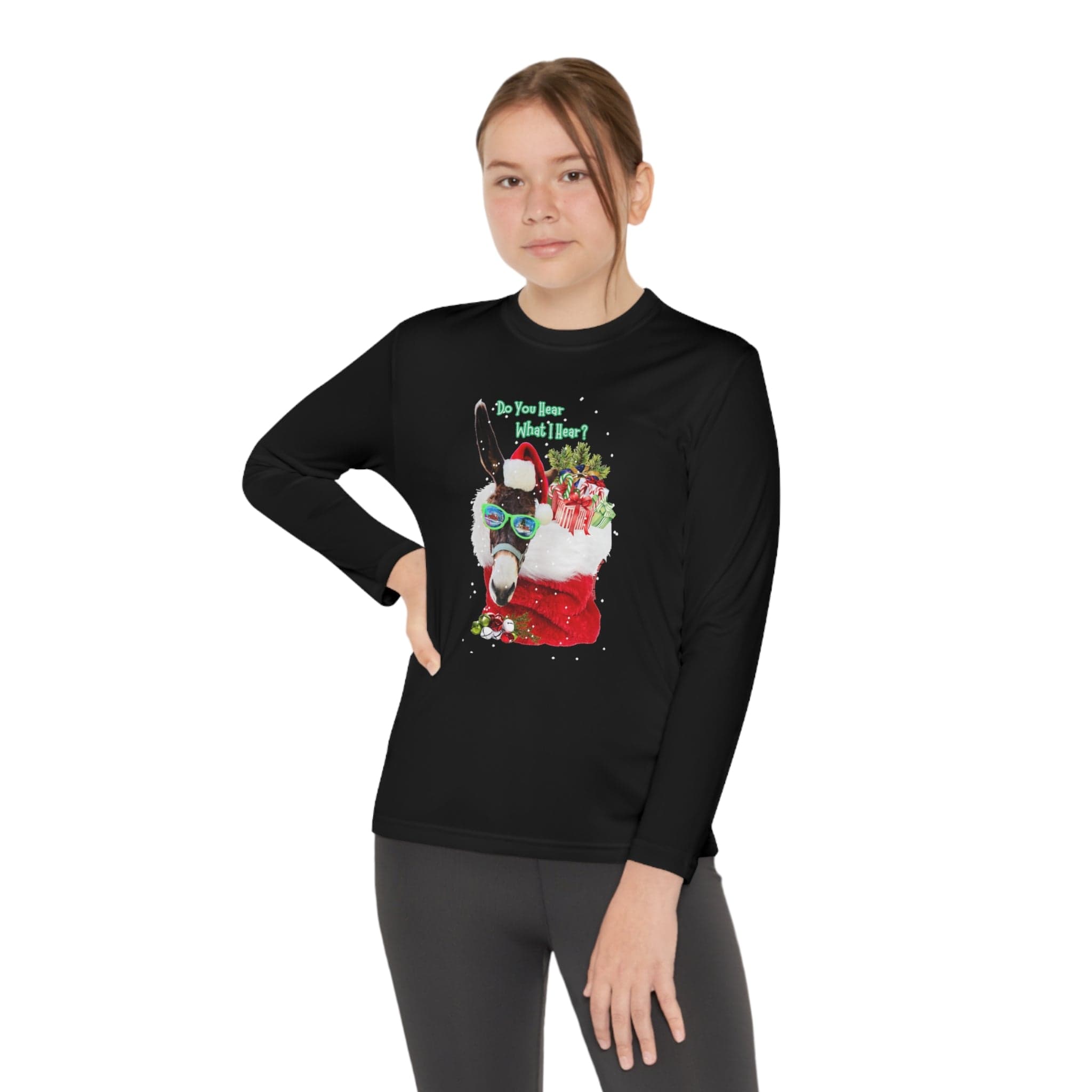 A person stands with one hand on their hip, donning the Urban SoulShine & co Do You Hear What I Hear Donkey Claus Long Sleeve Competitor Tee. This youth unisex tee, made from moisture-wicking polyester, features a festive Christmas llama and gift design that stands out against the plain white background.