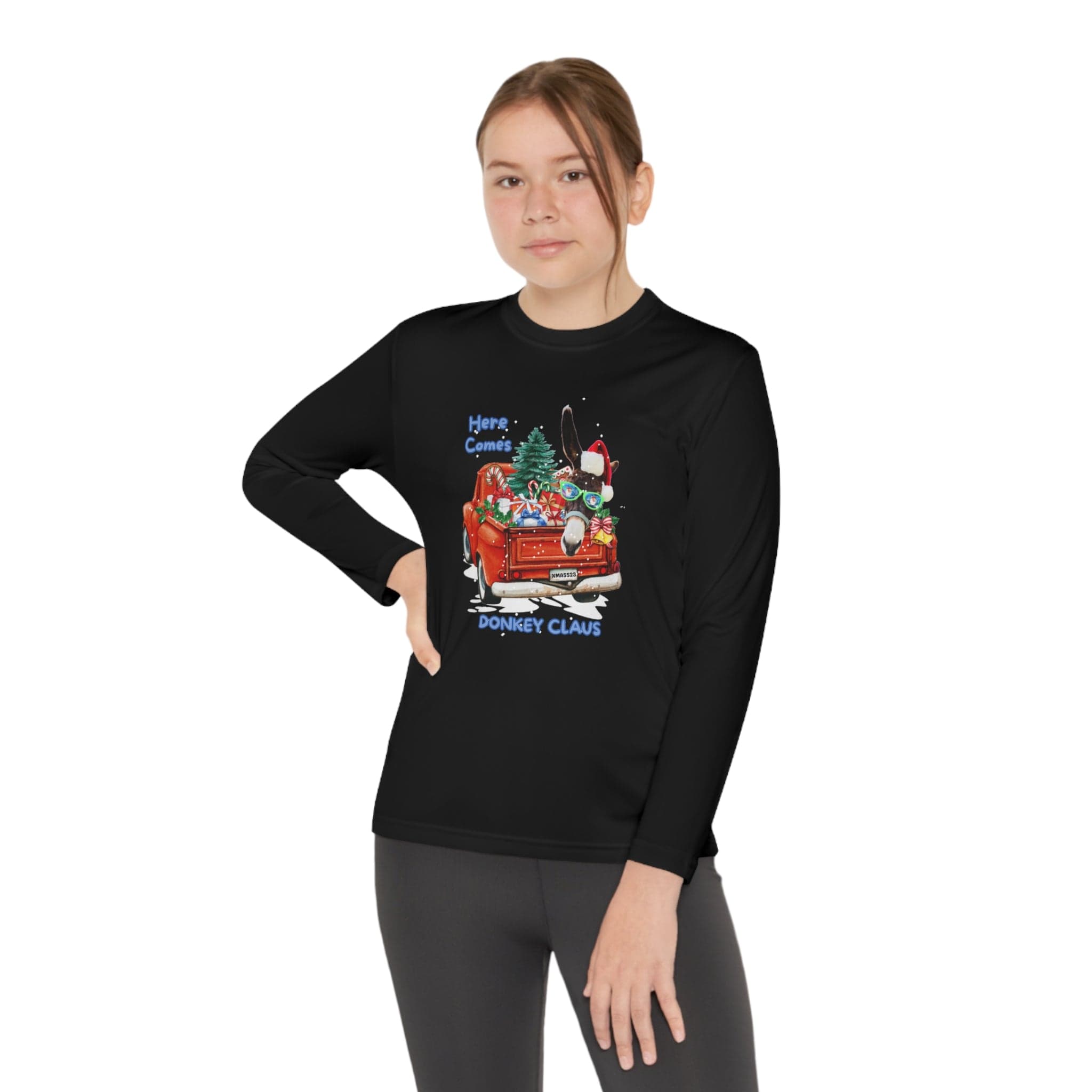 A young person is confidently wearing the Urban SoulShine & co "Here Comes Donkey Claus" Long Sleeve Competitor Tee for youth, made from moisture-wicking polyester in an athletic fit. The black shirt features a festive design with gifts, a Christmas tree, and a sleigh, complete with the text "Here Comes Donkey Claus." They stand with one hand on their hip against a neutral background.