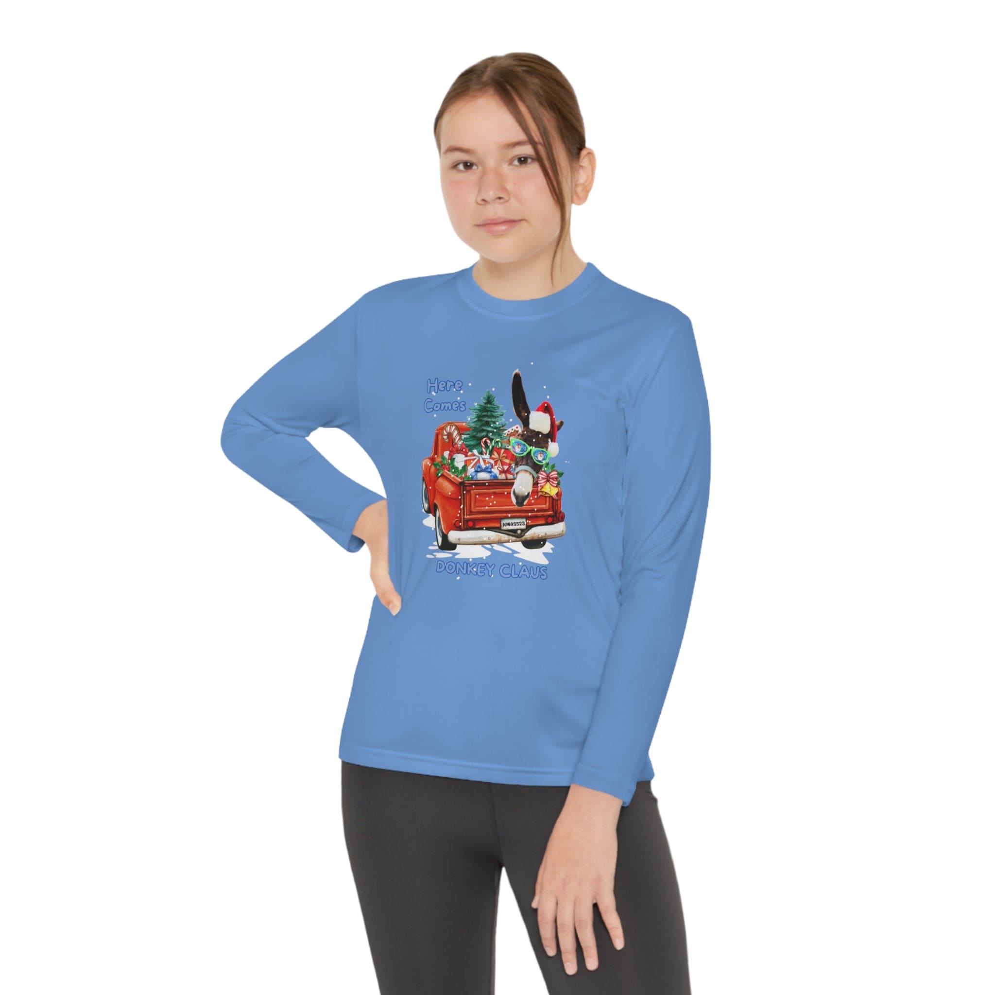 A person is wearing the Here Comes Donkey Claus Long Sleeve Competitor Tee (Youth Unisex) by Urban SoulShine & co. This vibrant blue tee has an athletic fit and features a festive design with a red truck carrying Christmas decorations. Made from moisture-wicking polyester, this Sport-Tek PosiCharge top ensures comfort as they stand with one hand on their hip against a plain white background.