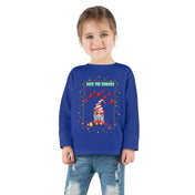 A young child with brown hair stands smiling in the Urban SoulShine & co Deck The Gnomes Christmas Toddler Long Sleeve Tee, crafted from 100% cotton in a festive blue. This charming shirt features a holiday gnome design along with the phrase "Deck the Gnomes," and is paired with light blue jeans to create an ideal ensemble for spreading cheer.