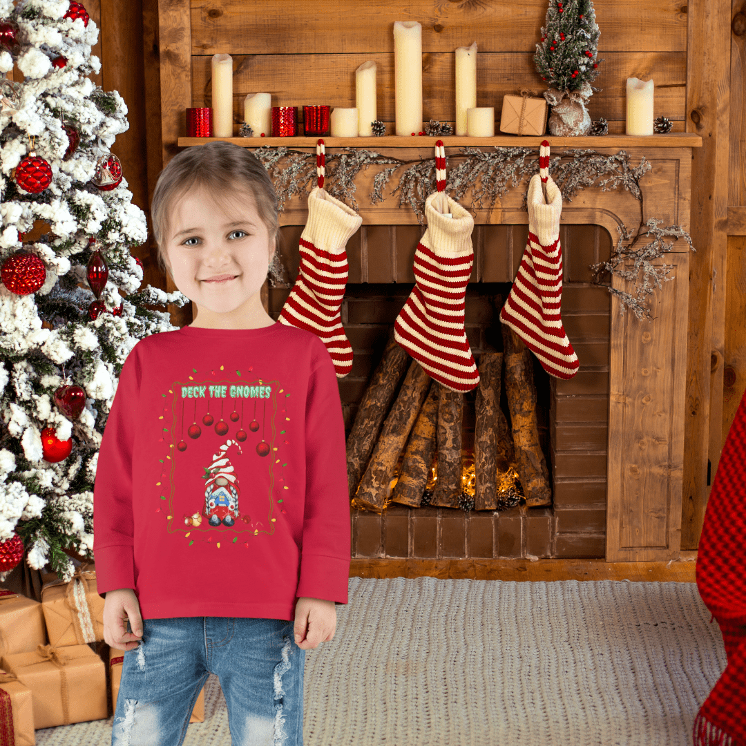 A young child in an Urban SoulShine & co Deck The Gnomes Christmas Toddler Long Sleeve Tee stands in front of a fireplace adorned with striped stockings and candles. On the left, a Christmas tree decorated with red and white ornaments is visible, with logs resting inside the hearth.