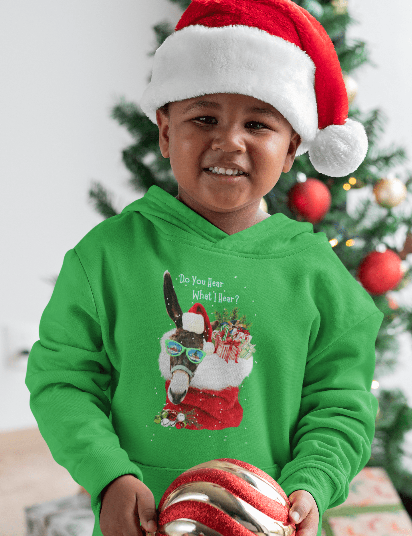 A young child in an Urban SoulShine & co's "Do You Hear What I Hear" Donkey Claus Hoodie (Youth Unisex) and a Santa hat beams while holding a large red and gold Christmas ornament. A decorated Christmas tree is visible in the background, enhancing the holiday cheer.