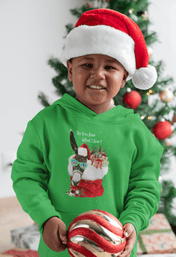 A young child in an Urban SoulShine & co's "Do You Hear What I Hear" Donkey Claus Hoodie (Youth Unisex) and a Santa hat beams while holding a large red and gold Christmas ornament. A decorated Christmas tree is visible in the background, enhancing the holiday cheer.