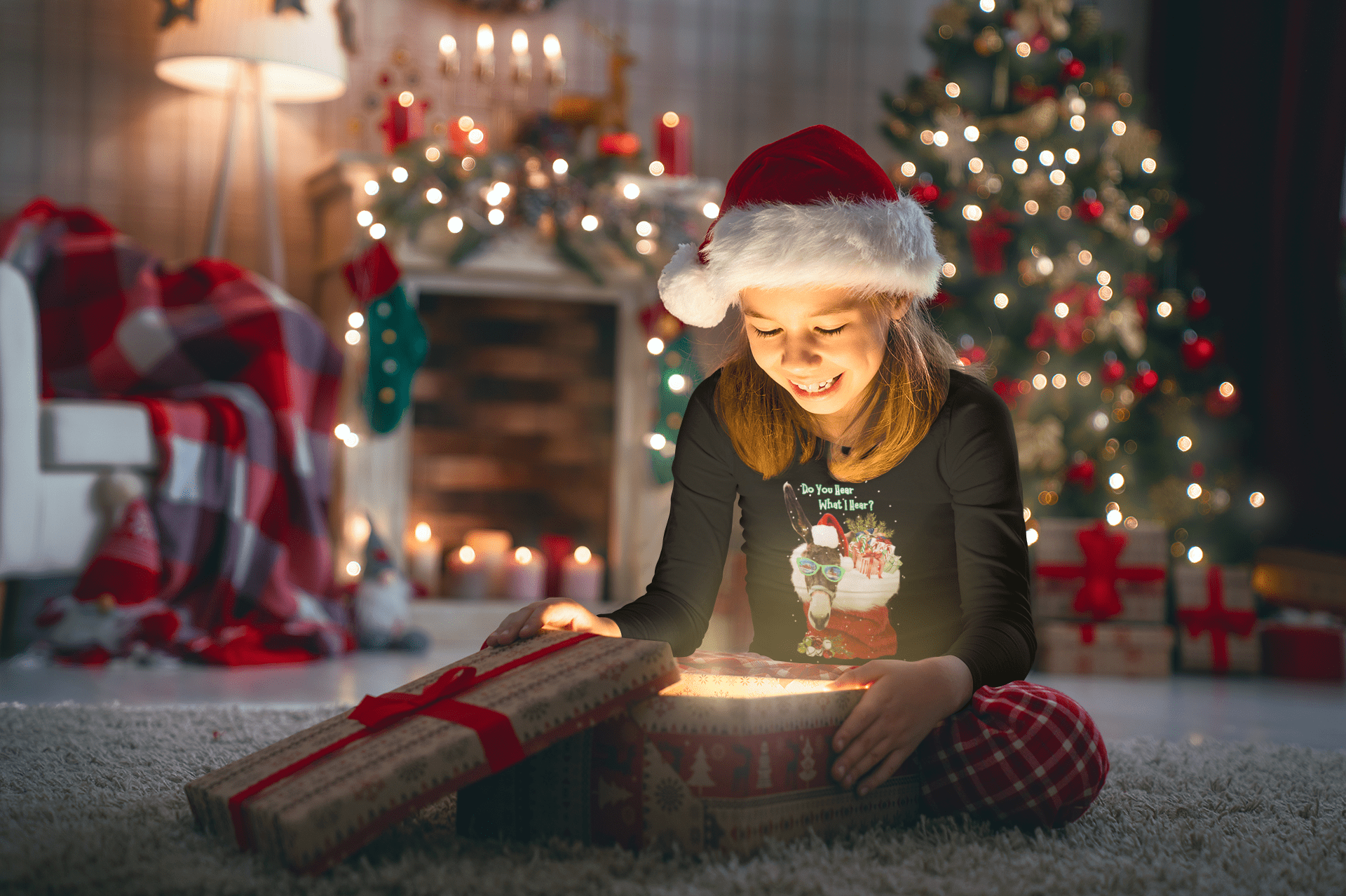 A child wearing a moisture-wicking Do You Hear What I Hear Donkey Claus Long Sleeve Competitor Tee (Youth Unisex) from Urban SoulShine & co sits on a carpet next to an open gift box, which emits a warm glow. In the background, a decorated Christmas tree stands near a lit fireplace with stockings hanging above it.