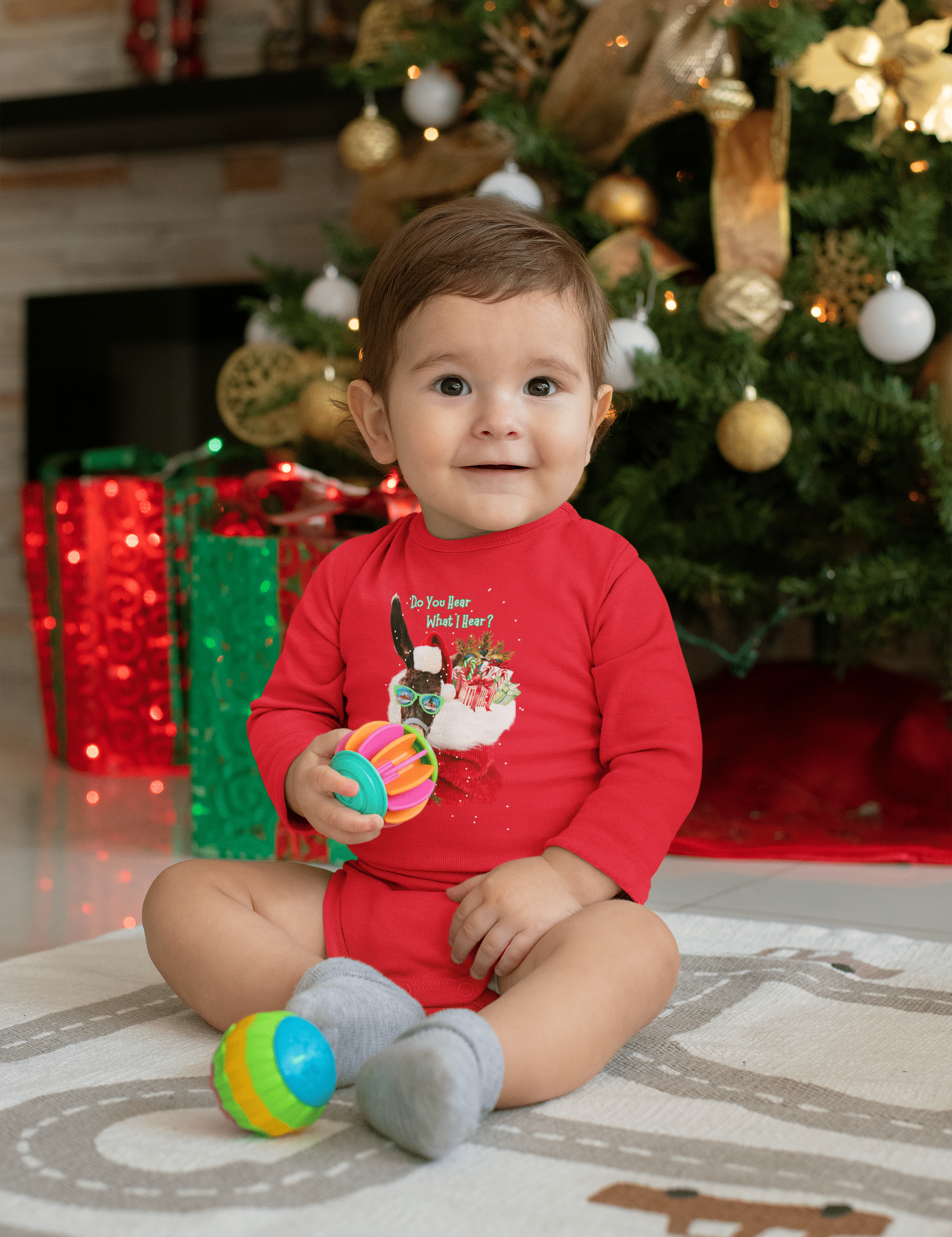 An infant wearing the "Do You Hear What I Hear Donkey Claus Long Sleeve Onesie" from Urban SoulShine & co sits on a rug, holding colorful balls. In the background, a beautifully decorated Christmas tree with lights and ornaments and wrapped gifts adds to the festive atmosphere. The baby is smiling and wears gray socks, enhancing the charm of this joyful scene.