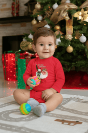 An infant wearing the "Do You Hear What I Hear Donkey Claus Long Sleeve Onesie" from Urban SoulShine & co sits on a rug, holding colorful balls. In the background, a beautifully decorated Christmas tree with lights and ornaments and wrapped gifts adds to the festive atmosphere. The baby is smiling and wears gray socks, enhancing the charm of this joyful scene.