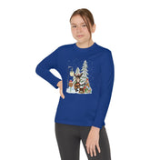 A person in a Donkey Claus Street Player Christmas Long Sleeve Competitor Tee from Urban SoulShine & co, featuring a festive design with a girl, a squirrel, and tall trees, strikes a pose next to a lamppost in the snow. The shirt's blue, moisture-wicking fabric is paired with dark pants as they stand with one hand on their hip against a plain white background.