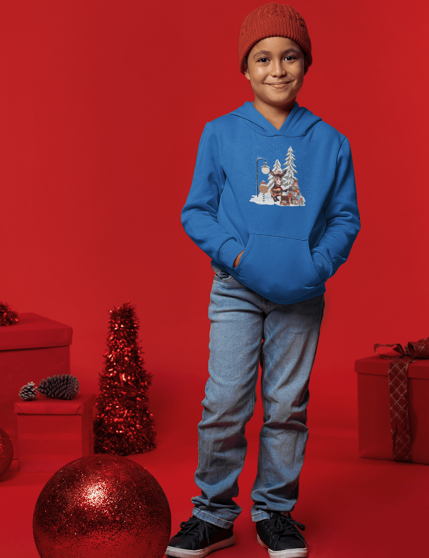 A child in a blue Donkey Claus Street Player Hoodie by Urban SoulShine & co stands against a red backdrop, wearing jeans, black sneakers, and a red beanie. They are surrounded by festive decorations, including a large bauble.