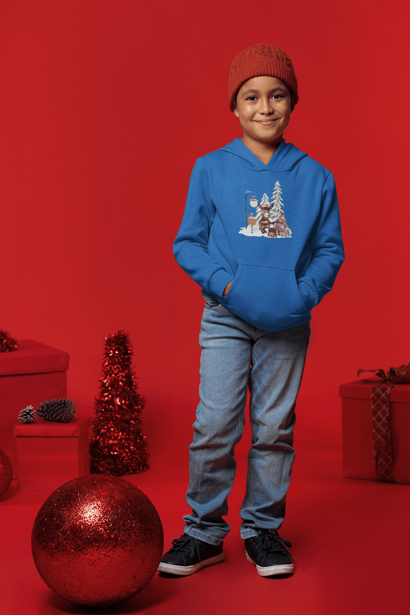 A child in a blue Donkey Claus Street Player Hoodie by Urban SoulShine & co stands against a red backdrop, wearing jeans, black sneakers, and a red beanie. They are surrounded by festive decorations, including a large bauble.