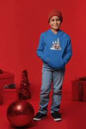 A child in a blue Donkey Claus Street Player Hoodie by Urban SoulShine & co stands against a red backdrop, wearing jeans, black sneakers, and a red beanie. They are surrounded by festive decorations, including a large bauble.