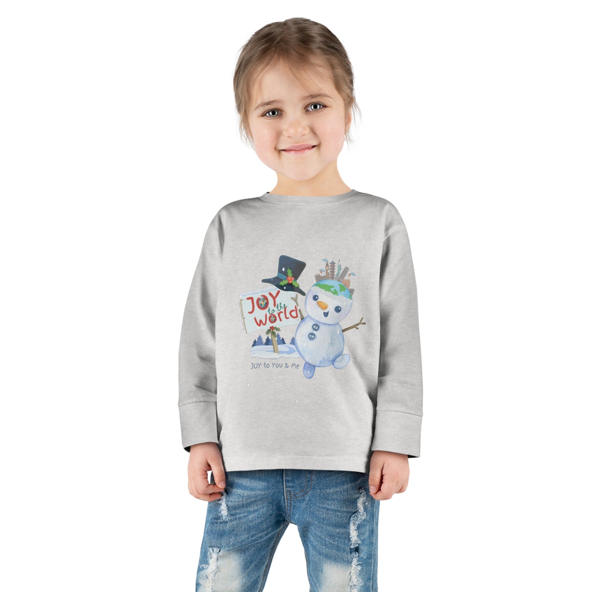 A young child is positioned against a white background, donning the Urban SoulShine & Co's "Joy To The World Snowman Christmas Toddler Long Sleeve Tee." With their hands casually tucked in their pockets, they are paired with classic blue jeans, showcasing the cheerful snowman design on the gray tee.