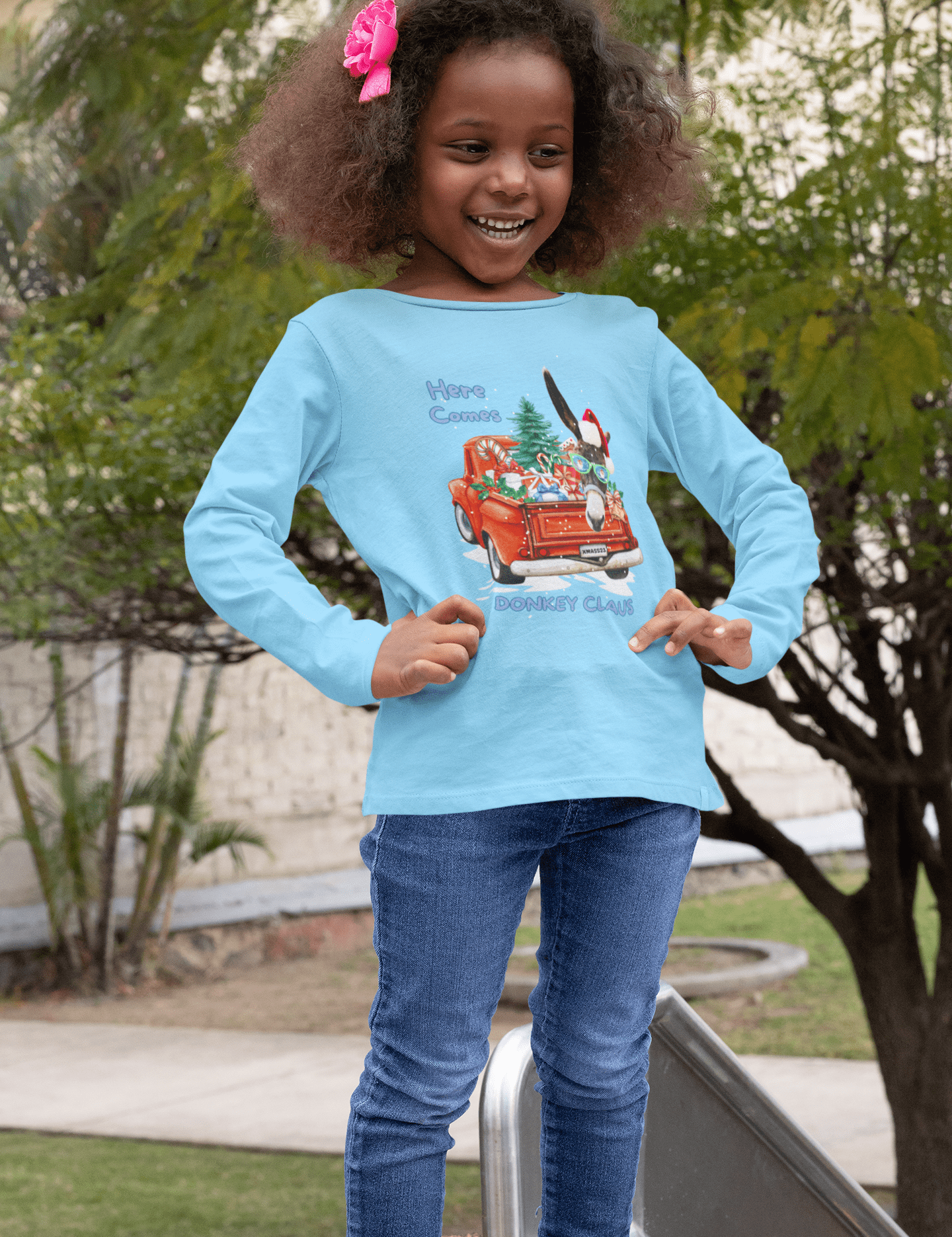 A young girl with curly hair, wearing a light blue Here Comes Donkey Claus Long Sleeve Competitor Tee by Urban SoulShine & co and jeans, stands smiling outdoors. Her shirt, made of moisture-wicking polyester, features a festive cartoon image of a red car with Christmas decorations. She is in a park area with trees and a slide visible behind her.