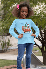 A young girl with curly hair, wearing a light blue Here Comes Donkey Claus Long Sleeve Competitor Tee by Urban SoulShine & co and jeans, stands smiling outdoors. Her shirt, made of moisture-wicking polyester, features a festive cartoon image of a red car with Christmas decorations. She is in a park area with trees and a slide visible behind her.