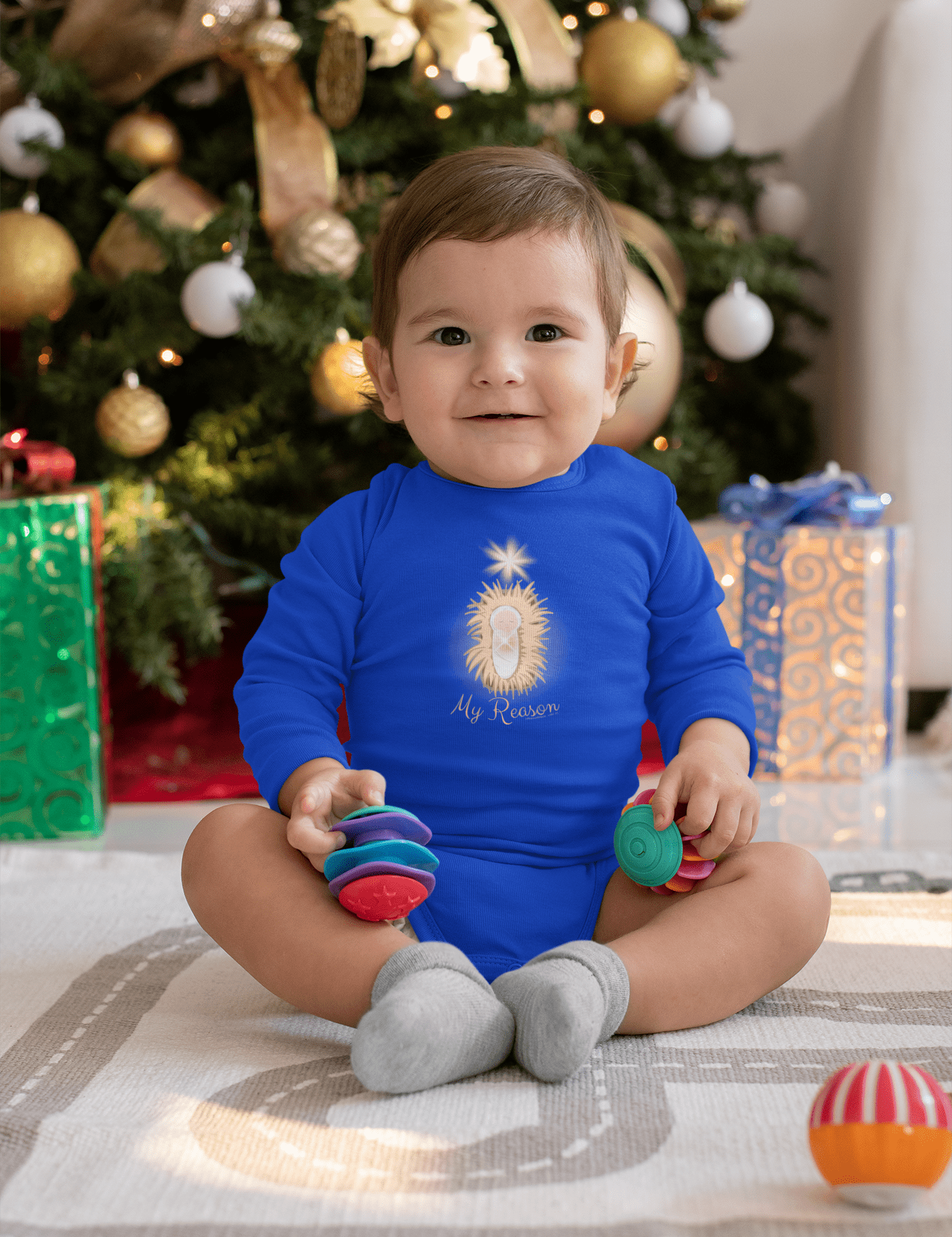 An infant wearing the "Jesus Is My Reason Long Sleeve Onesie Bodysuit" by Urban SoulShine & co is seated on a white blanket, playing with colorful toy rings. In the background, a decorated Christmas tree adorned with ornaments and surrounded by wrapped gifts adds to the festive ambiance.