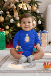 An infant wearing the "Jesus Is My Reason Long Sleeve Onesie Bodysuit" by Urban SoulShine & co is seated on a white blanket, playing with colorful toy rings. In the background, a decorated Christmas tree adorned with ornaments and surrounded by wrapped gifts adds to the festive ambiance.