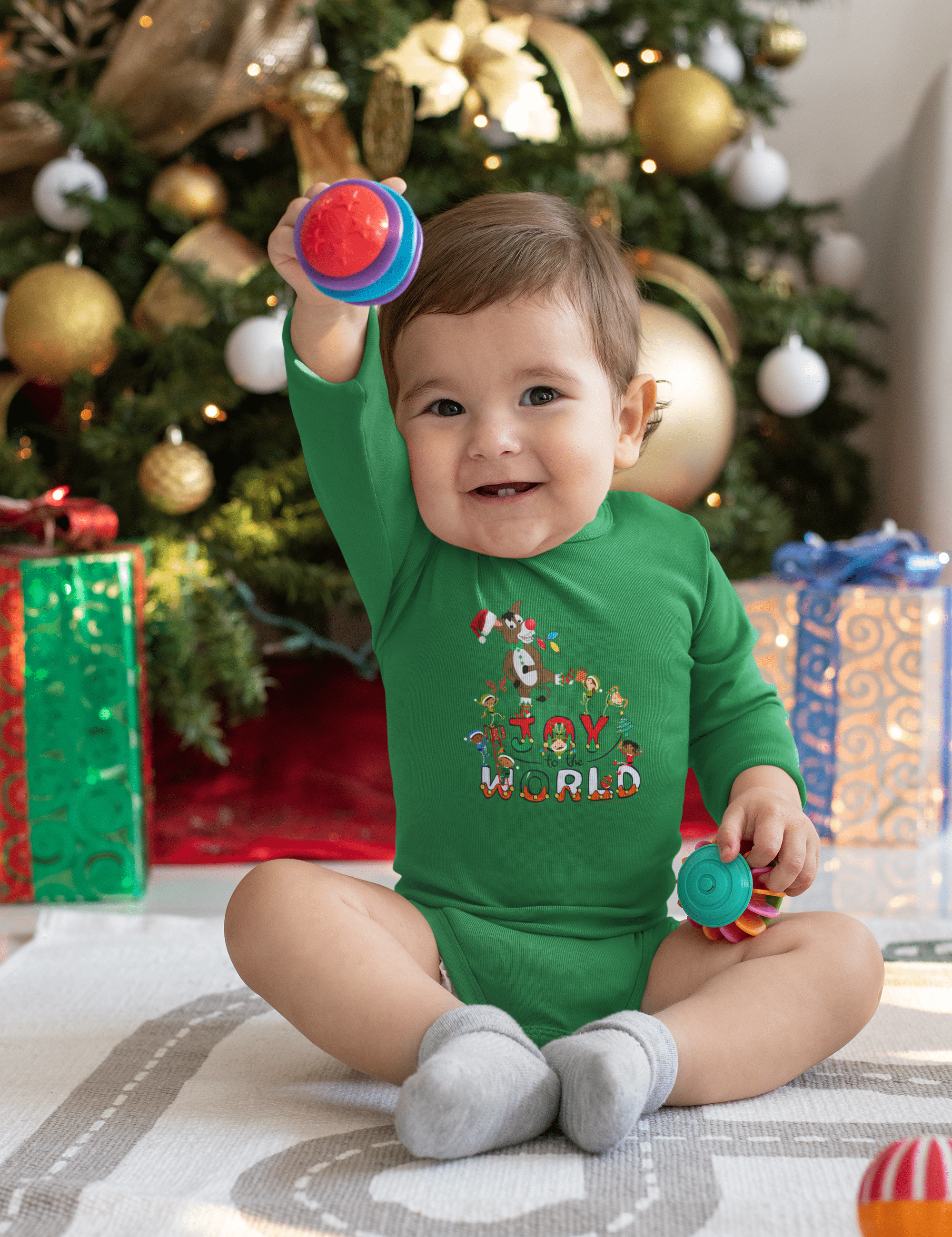 A baby wearing the Joy To The World Donkey Christmas Long Sleeve Onesie from Urban SoulShine & Co sits on a white blanket, holding colorful toys. In front of a decorated Christmas tree with wrapped presents beneath it, this cozy scene captures the essence of holiday joy.