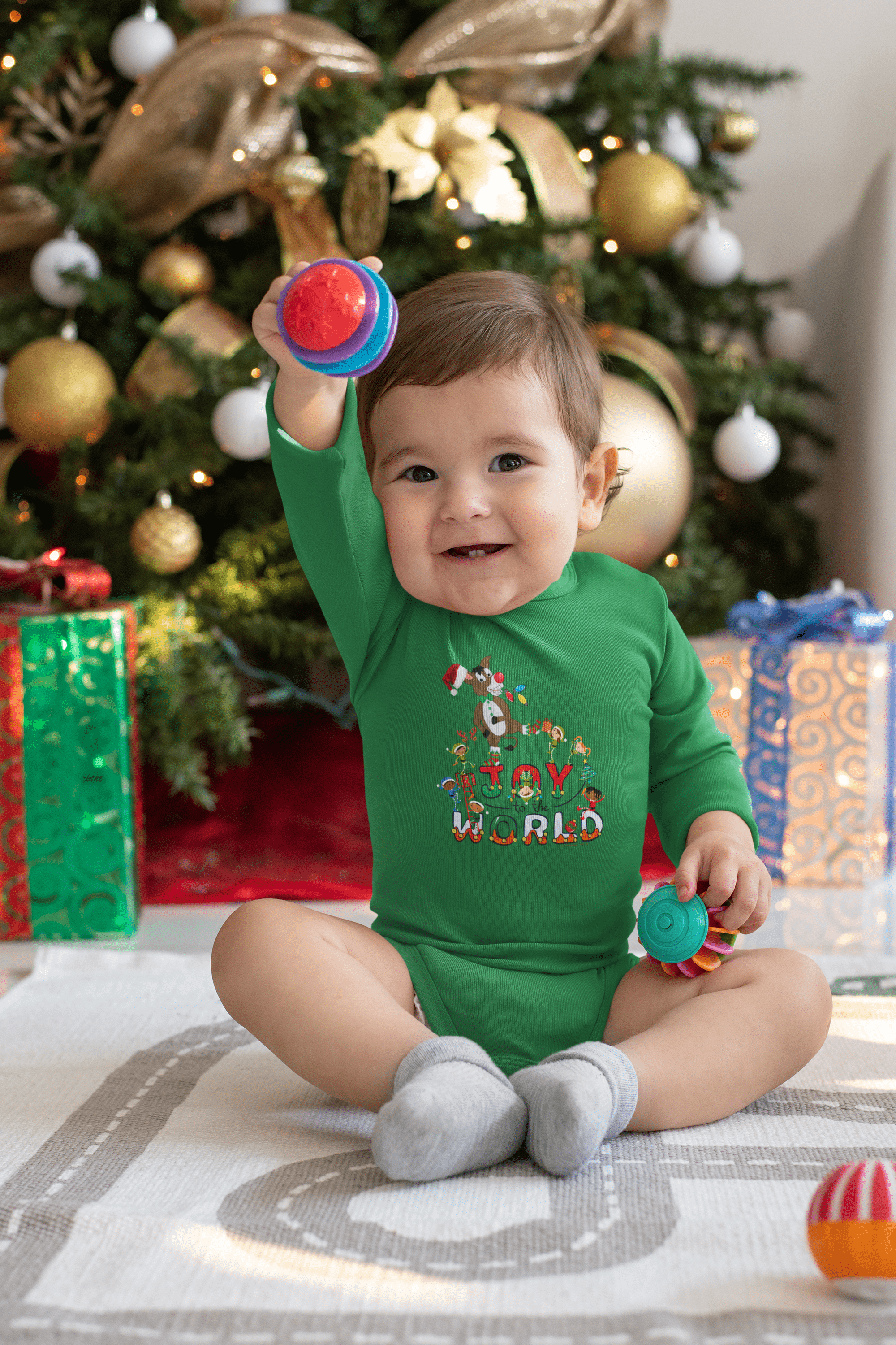 A baby wearing the Joy To The World Donkey Christmas Long Sleeve Onesie from Urban SoulShine & Co sits on a white blanket, holding colorful toys. In front of a decorated Christmas tree with wrapped presents beneath it, this cozy scene captures the essence of holiday joy.