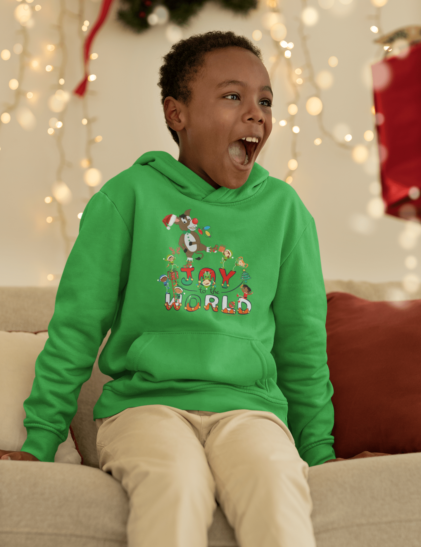 A child wearing the Joy To The World Donkey Claus Christmas Hoodie from Urban SoulShine & co is seated on a couch. The festive Donkey Claus design on the ultra-soft fleece youth hooded sweatshirt enhances the child's excitement, while holiday lights and decorations blur in the background.