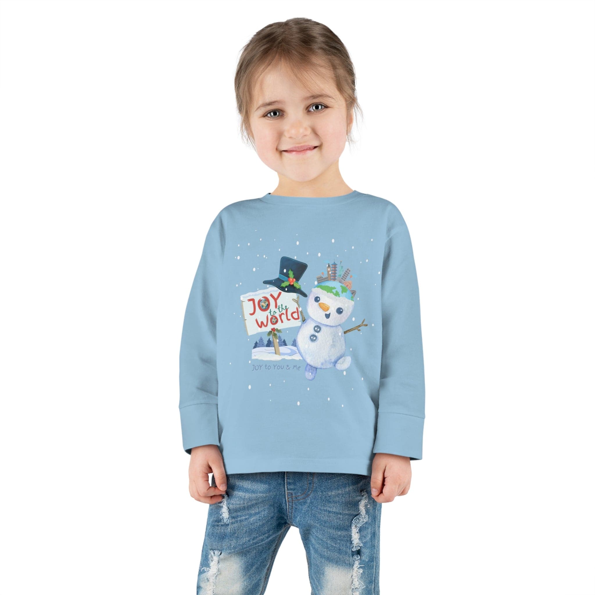 A young child stands smiling, wearing the Urban SoulShine & co Joy To The World Snowman Christmas Toddler Long Sleeve Tee. The light blue long-sleeve shirt, with its charming snowman design and "Joy to the World" text, pairs perfectly with their blue jeans and brown hair tied in a ponytail.