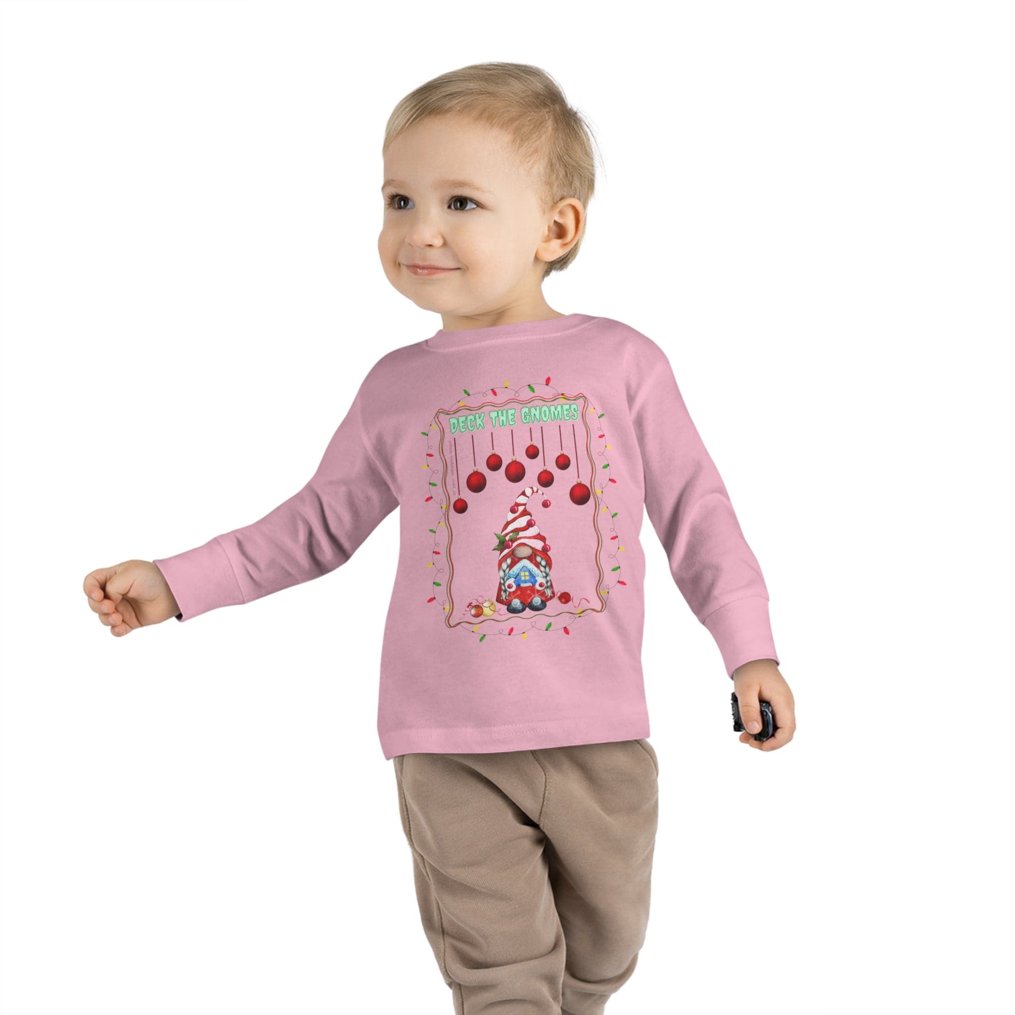 A small child in an Urban SoulShine & co "Deck The Gnomes Christmas Toddler Long Sleeve Tee" is smiling and holding a small object. The 100% cotton pink shirt features a holiday gnome design with red ornaments and "Deck the Gnomes" text, pairing perfectly with tan pants against a plain white background.