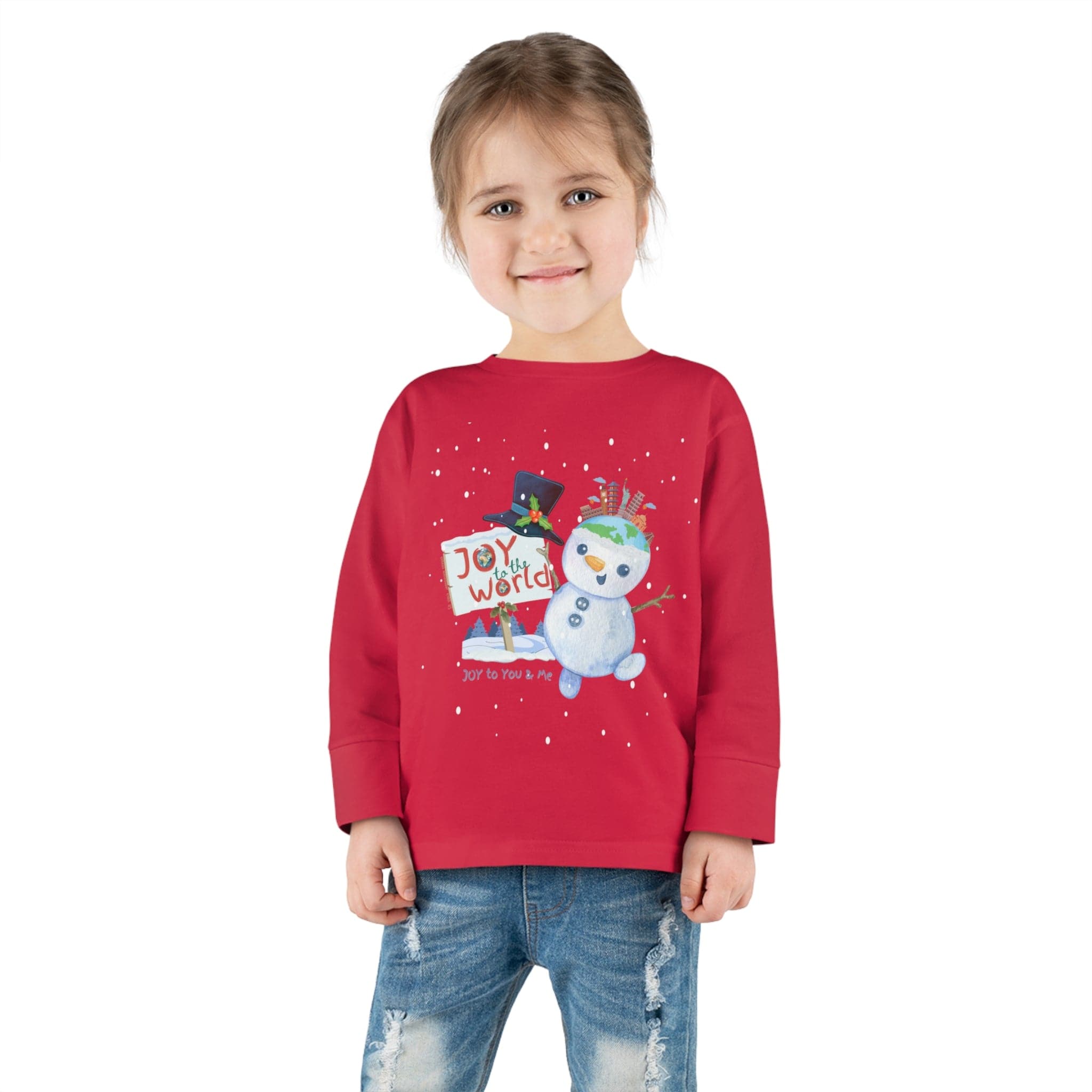 A young child stands smiling, dressed in the "Joy To The World Snowman Christmas Toddler Long Sleeve Tee" by Urban SoulShine & co. The vibrant red shirt with its snowman design and joyful message pairs perfectly with blue jeans, while their simply styled hair adds to the festive charm ideal for any Christmas occasion.