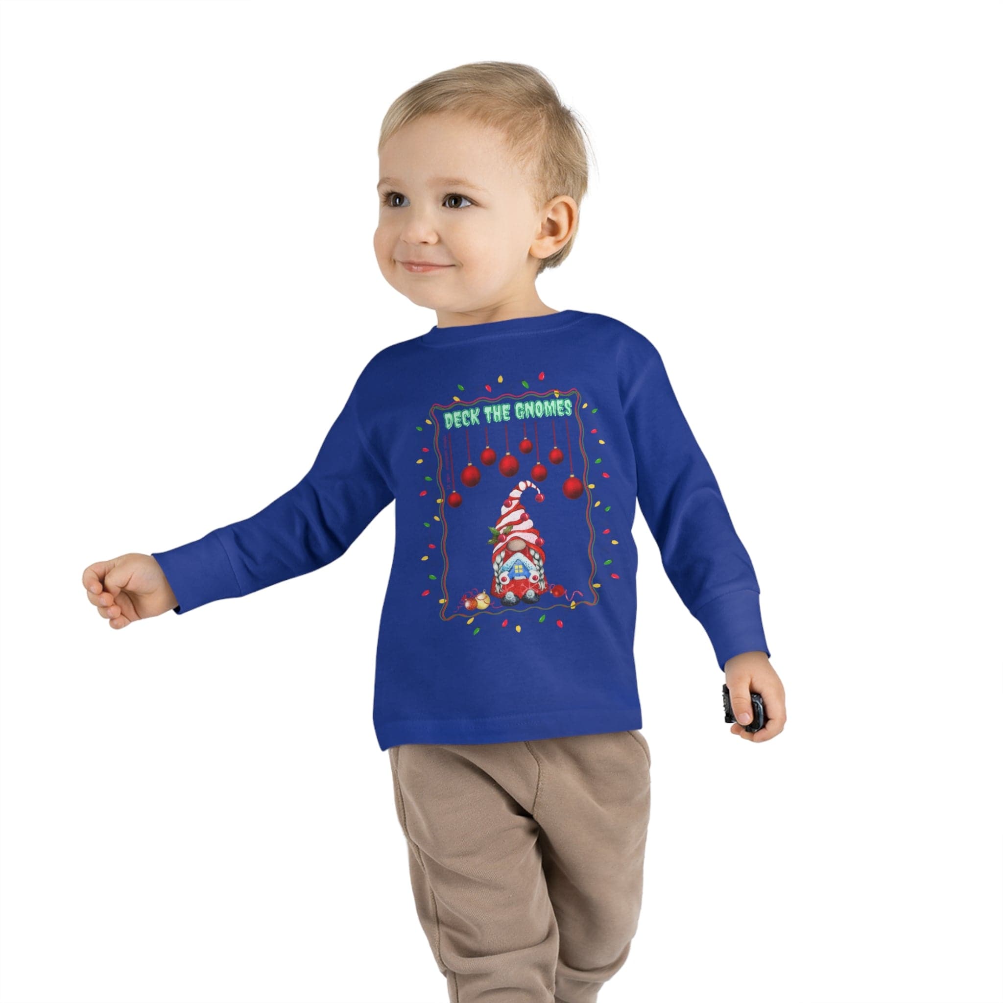 A young child in a blue "Deck The Gnomes Christmas Toddler Long Sleeve Tee" from Urban SoulShine & co, adorned with a festive holiday gnome design, walks forward with a small black object in hand. The shirt, made from 100% cotton, complements the child's light hair and smiling demeanor.