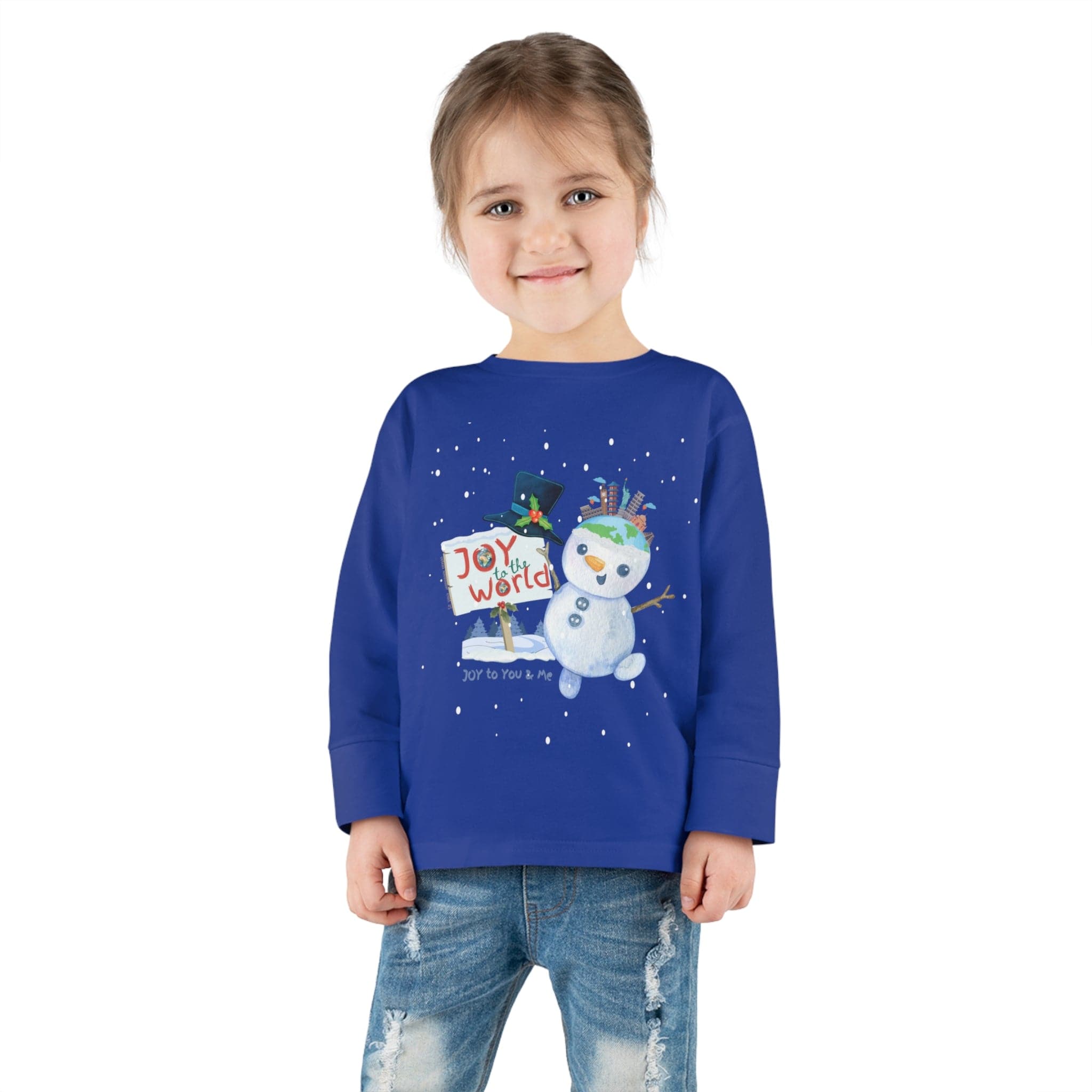 A young child with long hair is beaming, dressed in the Urban SoulShine & co Joy To The World Snowman Christmas Toddler Long Sleeve Tee. Paired with jeans, the child stands against a plain white background.