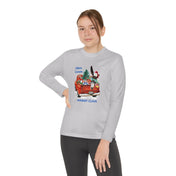 Dressed in Urban SoulShine & co's "Here Comes Donkey Claus Long Sleeve Competitor Tee (Youth Unisex)," a person confidently stands with one hand on their hip against a white background. This athletic fit gray tee features a festive design depicting a red vehicle carrying colorful gifts and a tree. Made from moisture-wicking polyester, it includes the playful text, "Here Comes Donkey Claus.