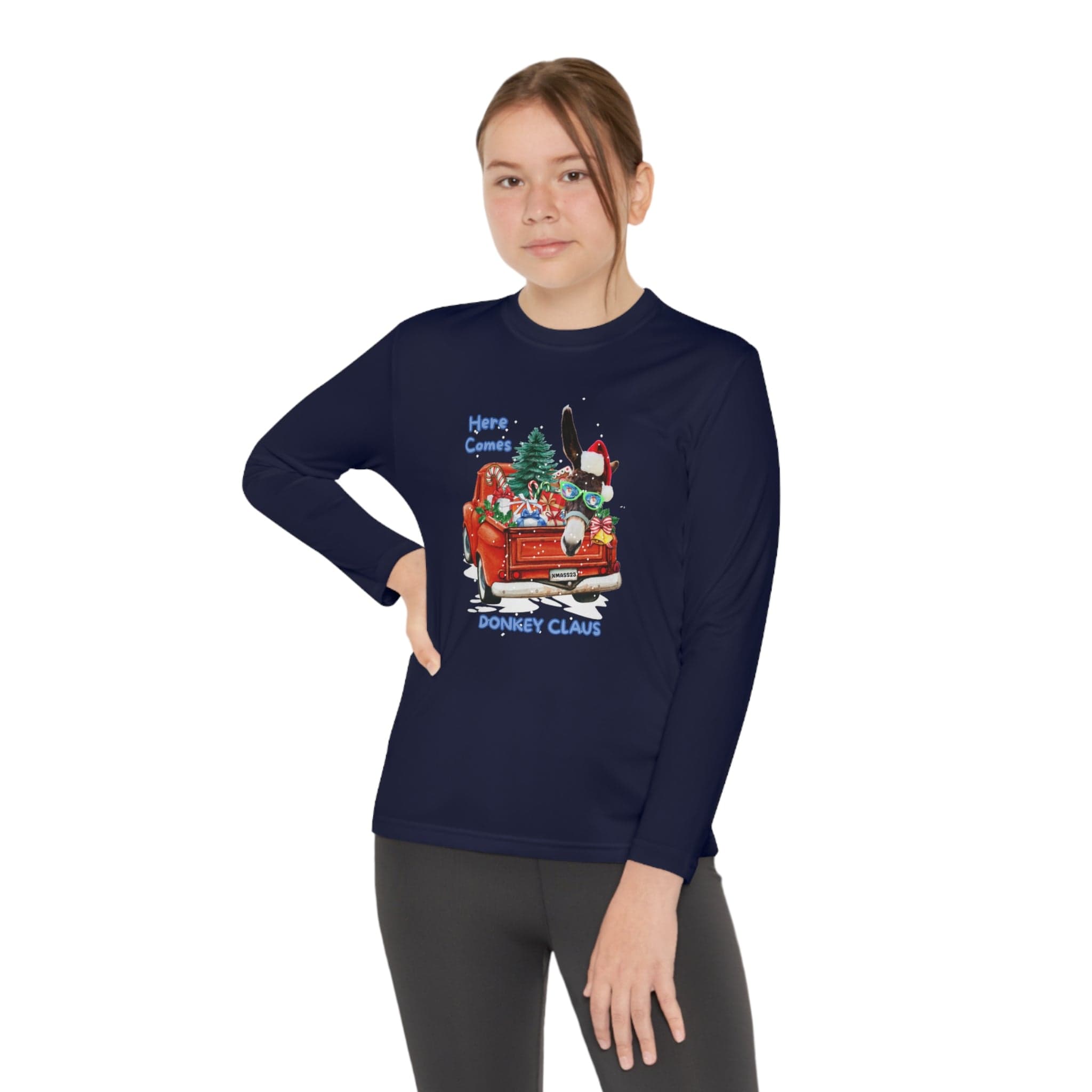 A person poses with one hand on their hip, displaying the athletic fit of a youth unisex Here Comes Donkey Claus Long Sleeve Competitor Tee by Urban SoulShine & co. The festive design includes a donkey driving a red car with gifts, adorned by a Christmas tree and the text "Here Comes Donkey Claus." Their hair is neatly tied in a ponytail.