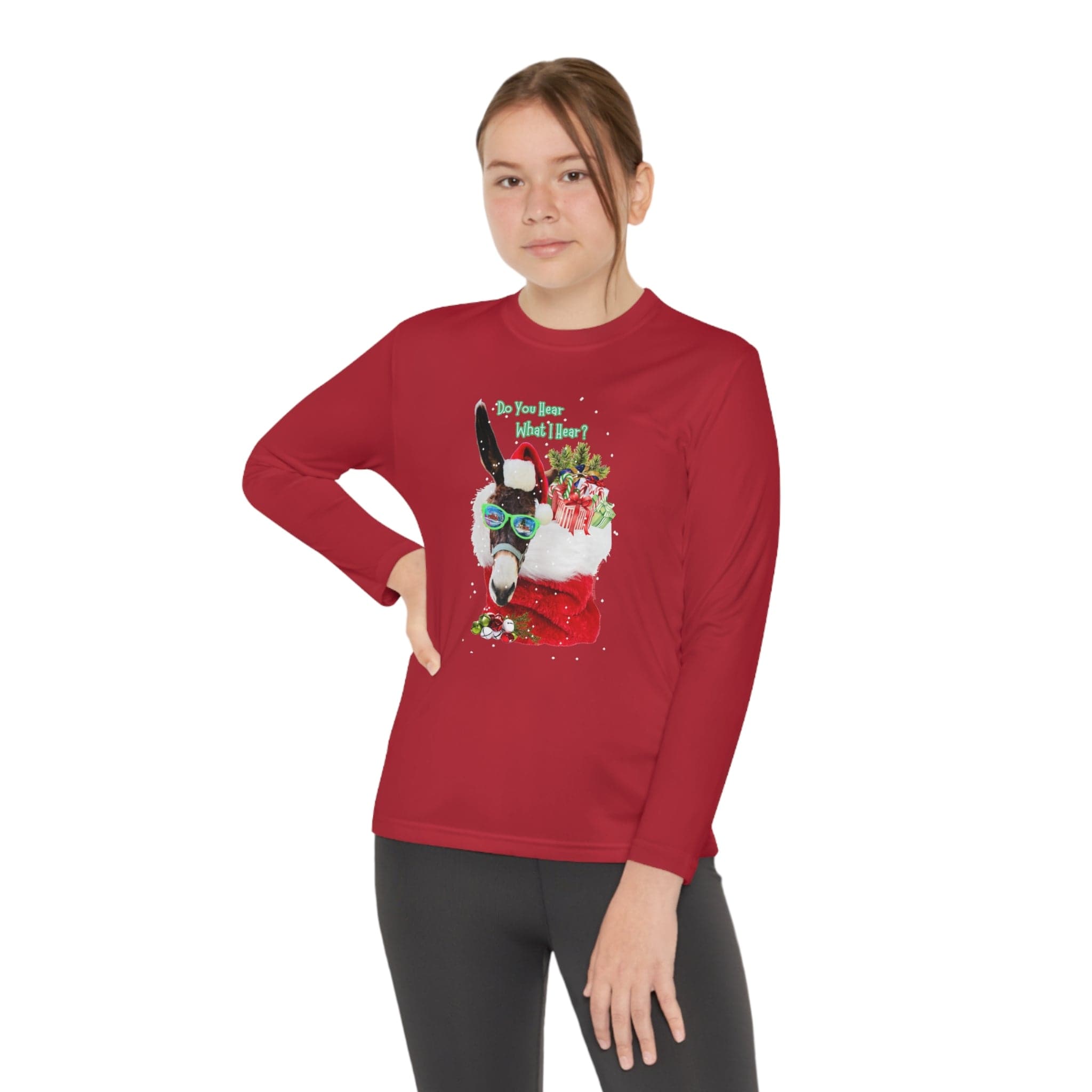 A person stands with one hand on their hip, dressed in an Urban SoulShine & co "Do You Hear What I Hear Donkey Claus" Long Sleeve Competitor Tee (Youth Unisex) in red, adorned with a festive design featuring Donkey Claus and presents. The moisture-wicking polyester shirt is complemented by black pants against a plain white background.