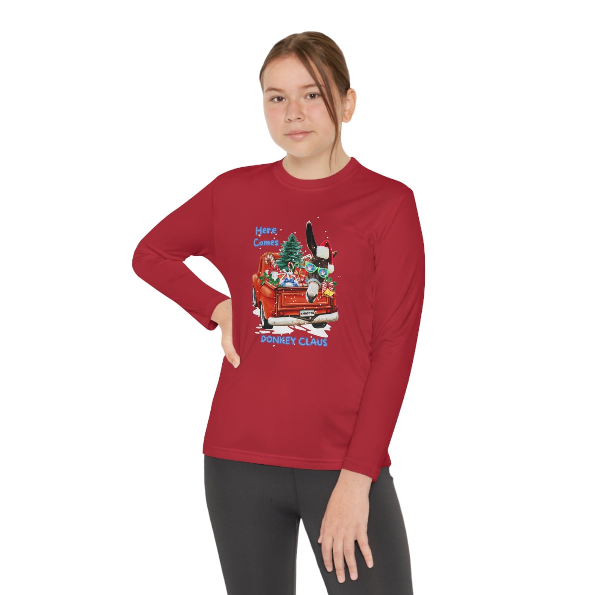 A person wearing a Here Comes Donkey Claus Long Sleeve Competitor Tee (Youth Unisex) by Urban SoulShine & co, featuring an athletic fit, red color, and a festive cartoonish donkey dressed as Santa in a red car graphic, poses confidently with one hand on their hip. The moisture-wicking polyester design pairs perfectly with their black pants.